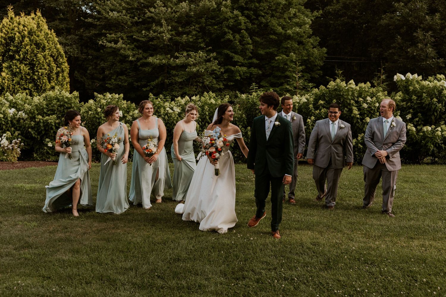 Wedding party gathered on lawn with bridesmaids in sage green dresses.
