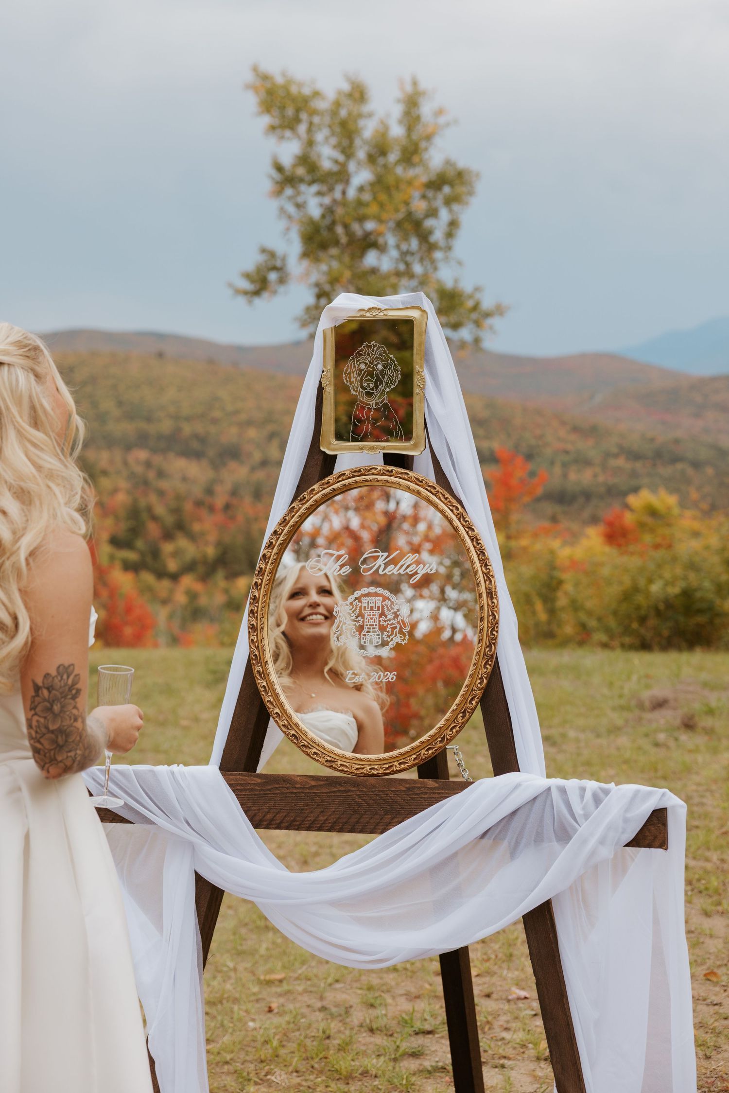 A bride views her reflection in an ornate gold oval mirror on an easel amid autumn mountain scenery.