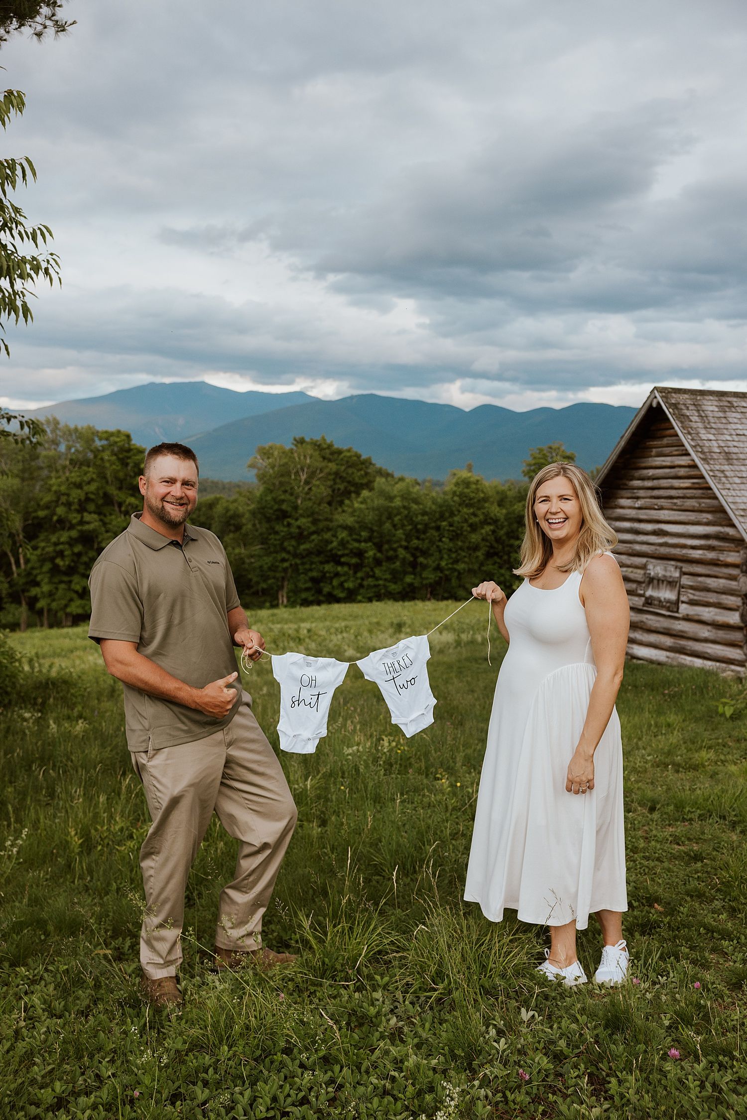 New Hampshire couple holding baby onesies on a clothesline in a mountain meadow, announcing a twin pregnancy outdoors.