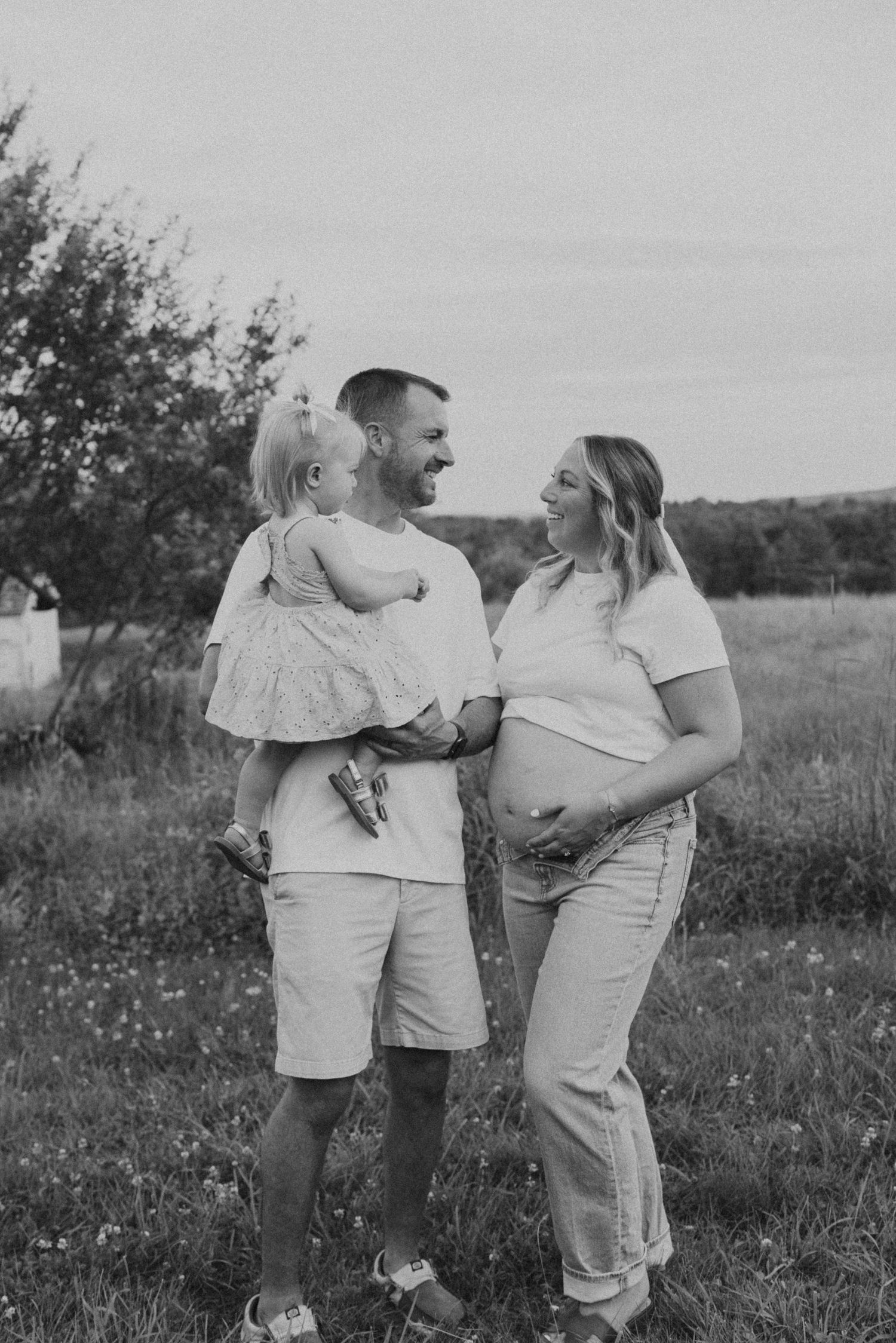Black and white photo of pregnant woman and partner smiling at toddler in an outdoor field setting.