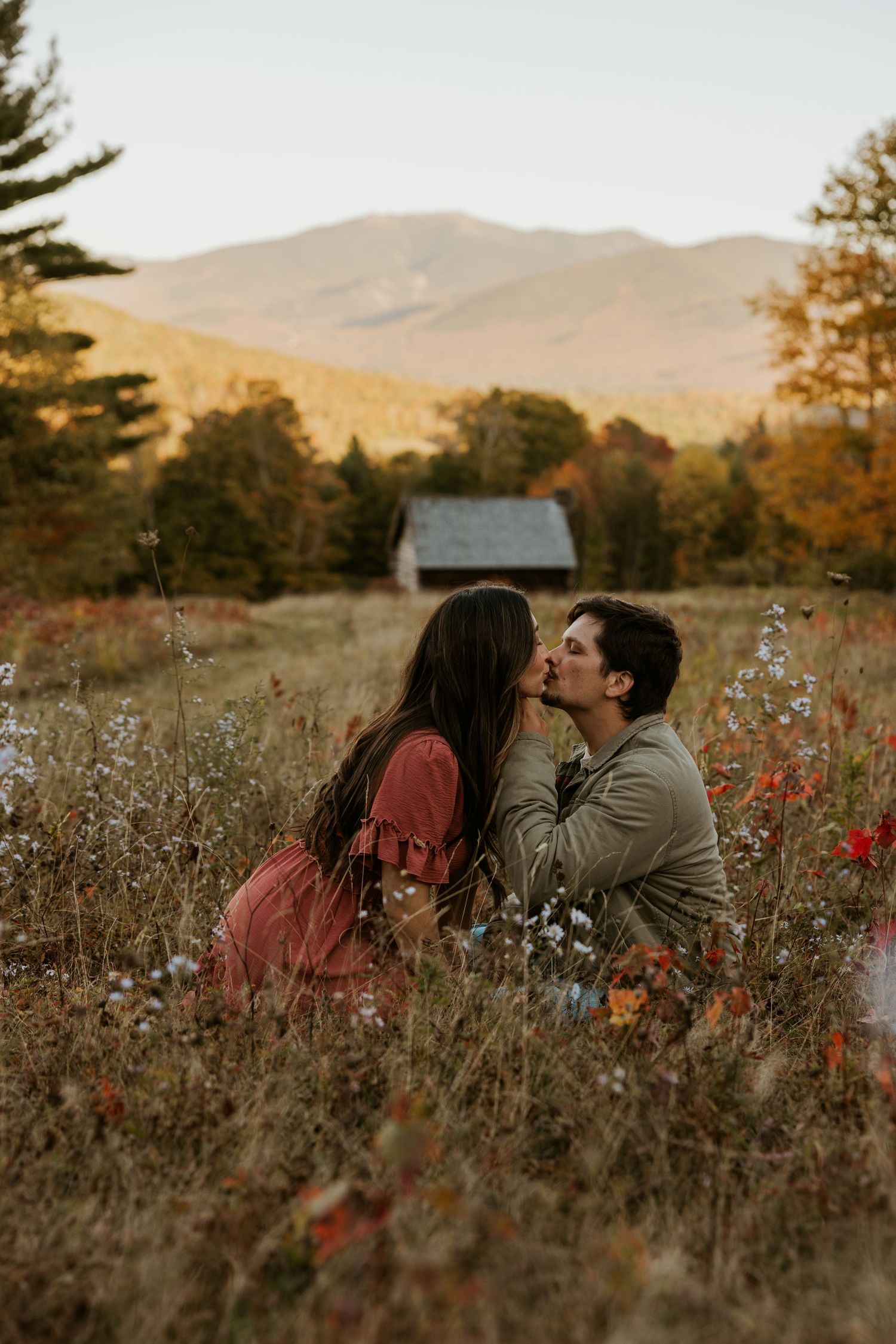 Romantic couple kissing in fall meadow with colorful foliage and mountain views behind them.