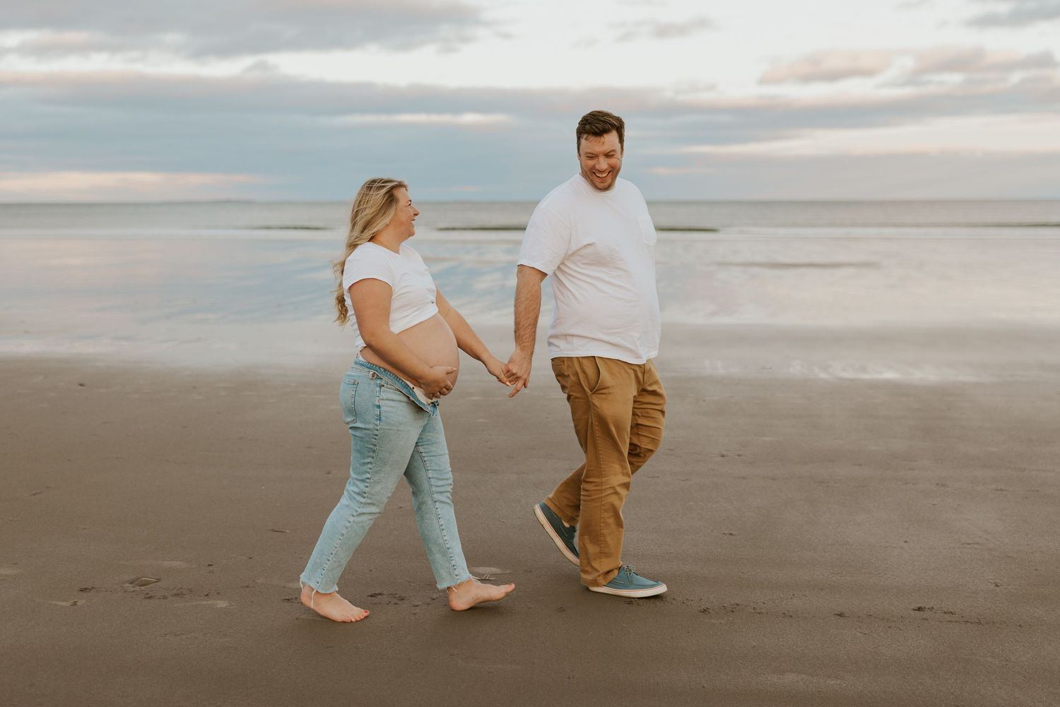 Smiling pregnant couple walking hand in hand barefoot on a serene beach.