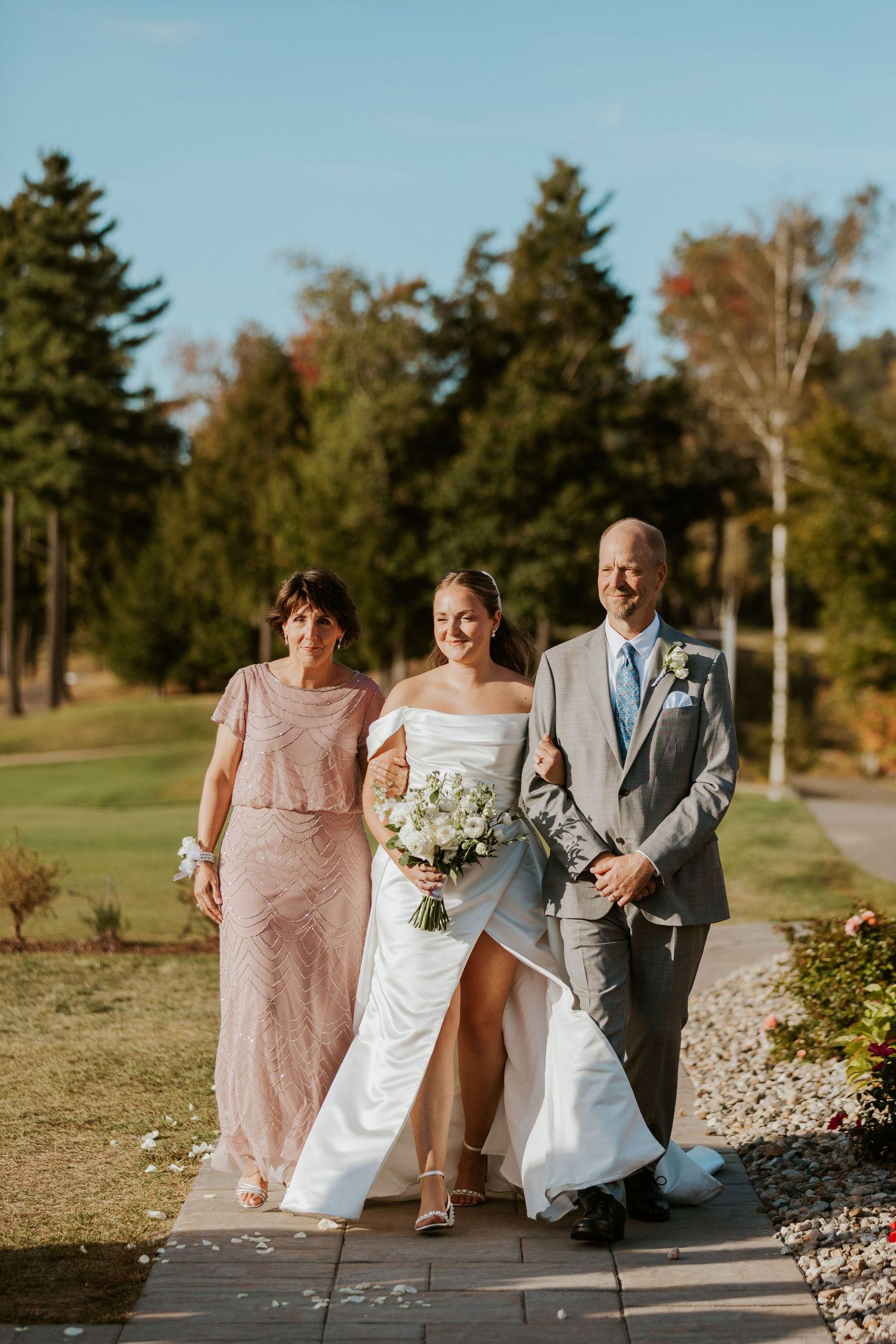 Bride in white off-shoulder gown walking down aisle with parents on sunny wedding day.