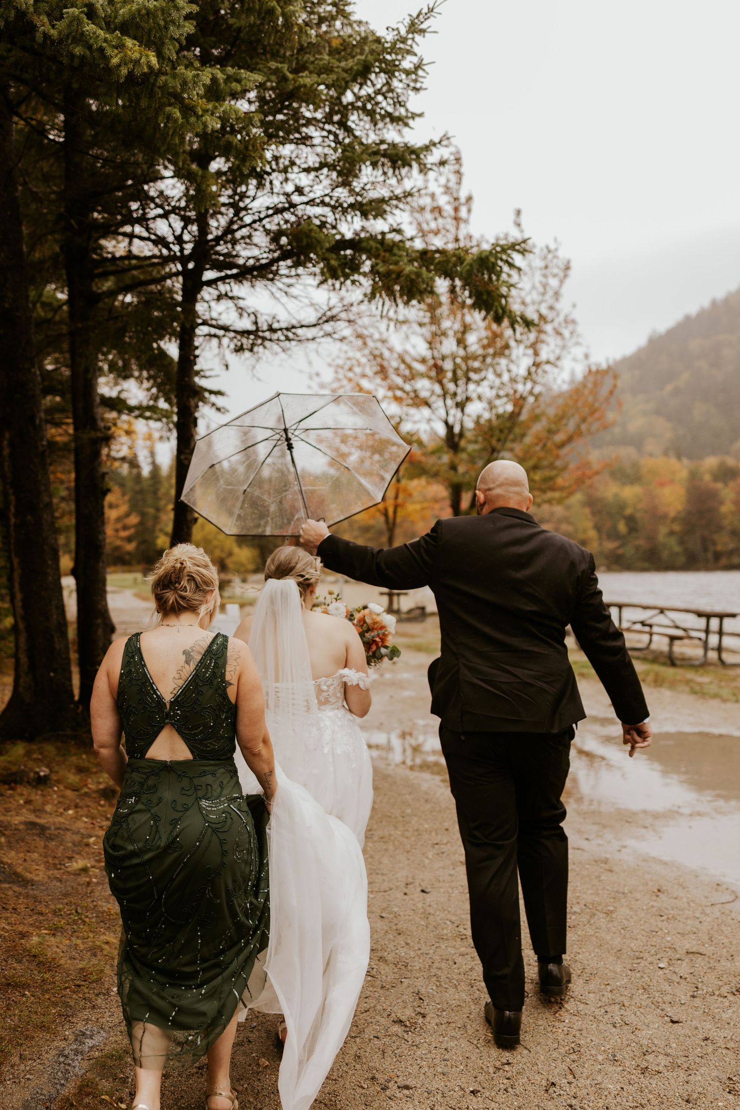 Bride walks with parents along a rainy autumn path toward a misty lake, umbrella held overhead.