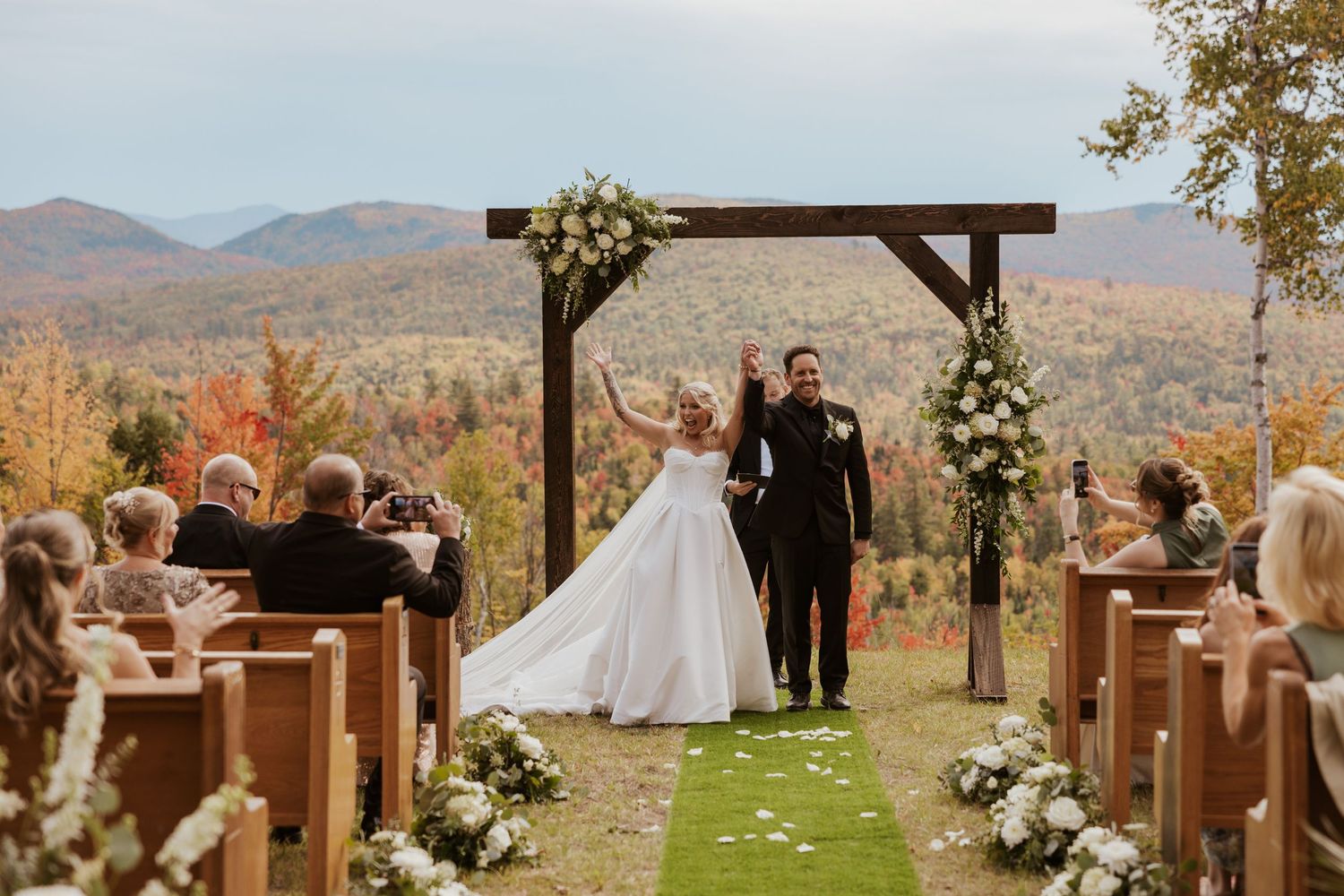 Bride and groom walk down outdoor aisle under wooden arch with autumn mountain backdrop at fall wedding ceremony.