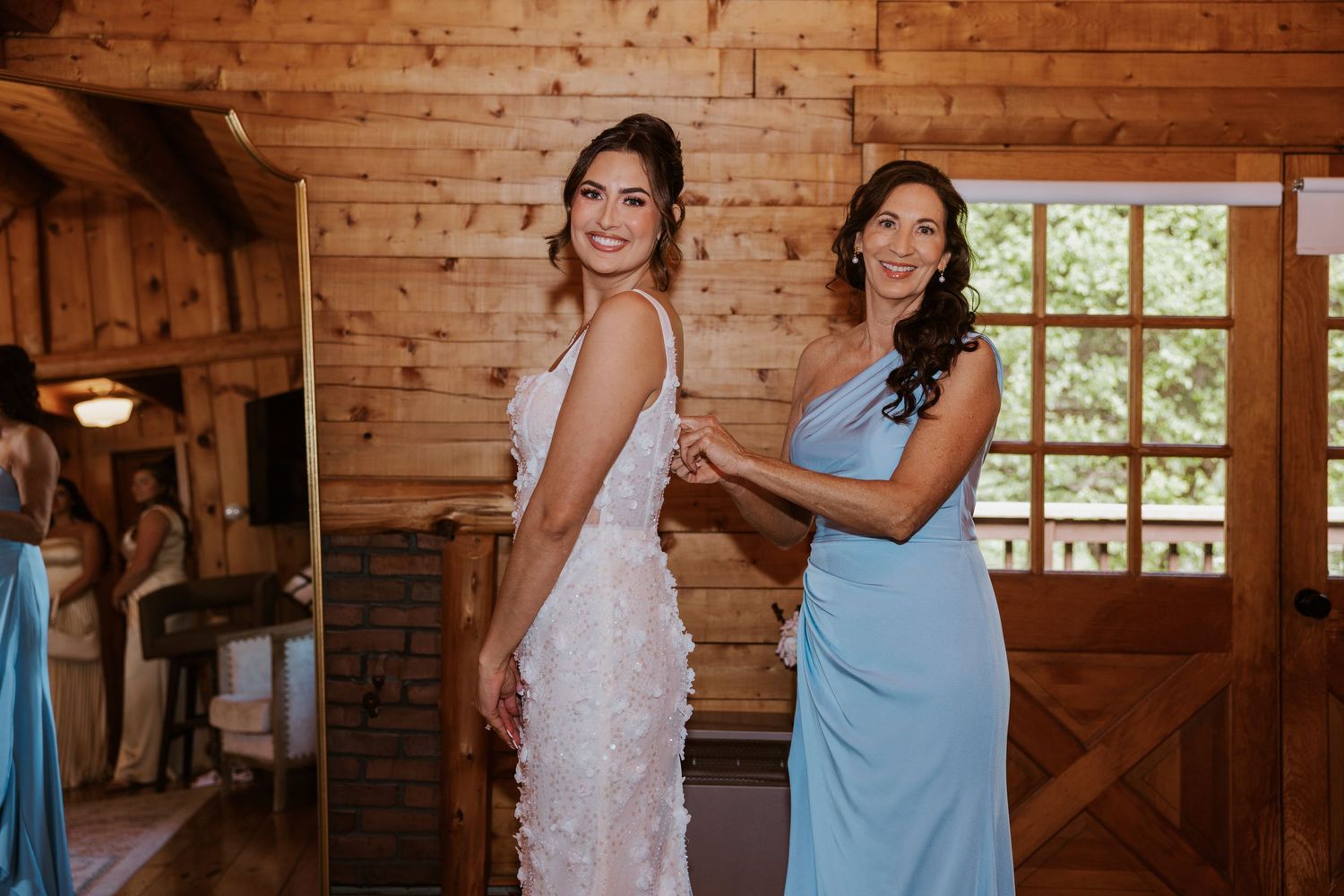 Bridesmaid in blue dress helping bride in white lace gown get dressed inside a rustic wooden barn venue.