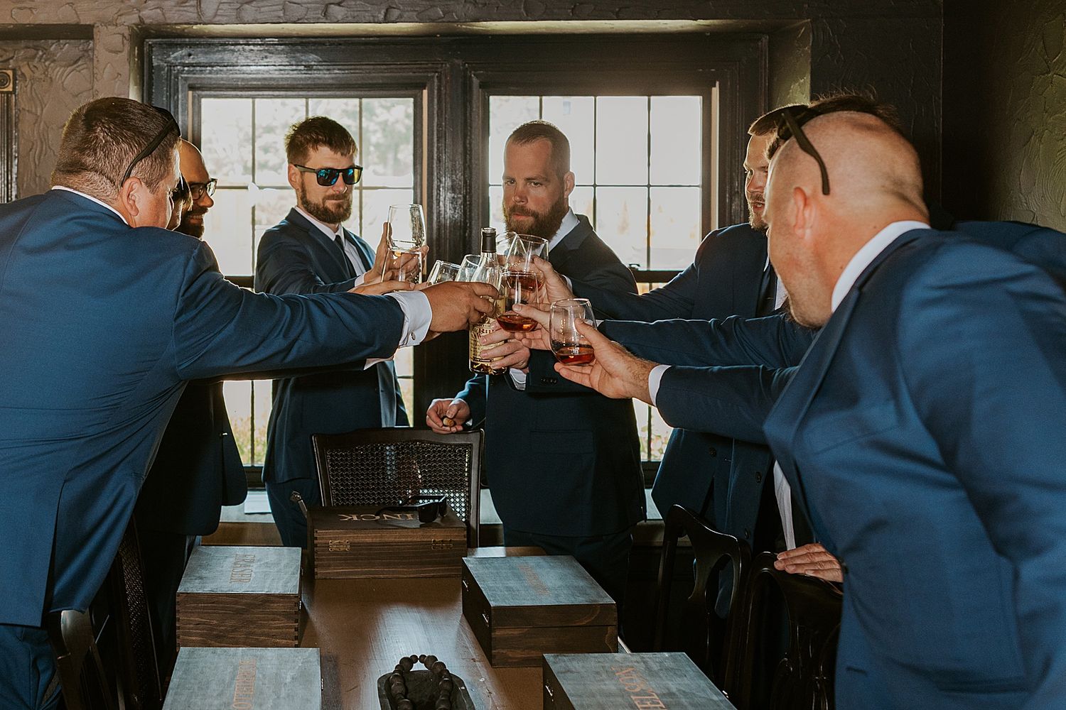 Five groomsmen in blue suits toasting with whiskey glasses at a wooden bar before a New Hampshire wedding ceremony