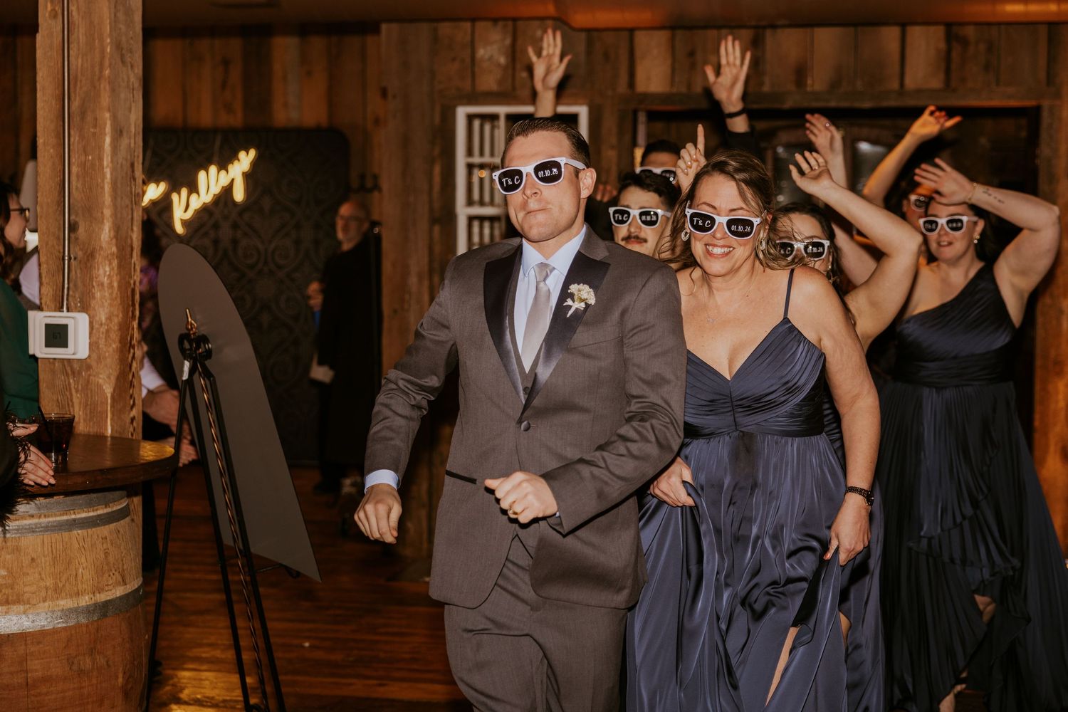 Wedding party in sunglasses celebrating at rustic barn reception entrance, bridesmaids in navy dresses cheering.