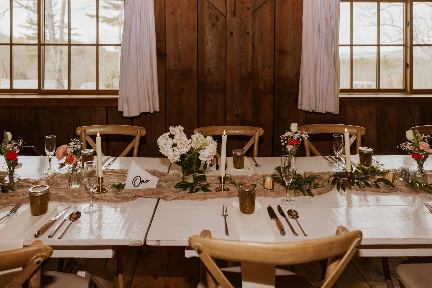 Long wooden tables set for a rustic barn wedding reception with natural decor and white linens.
