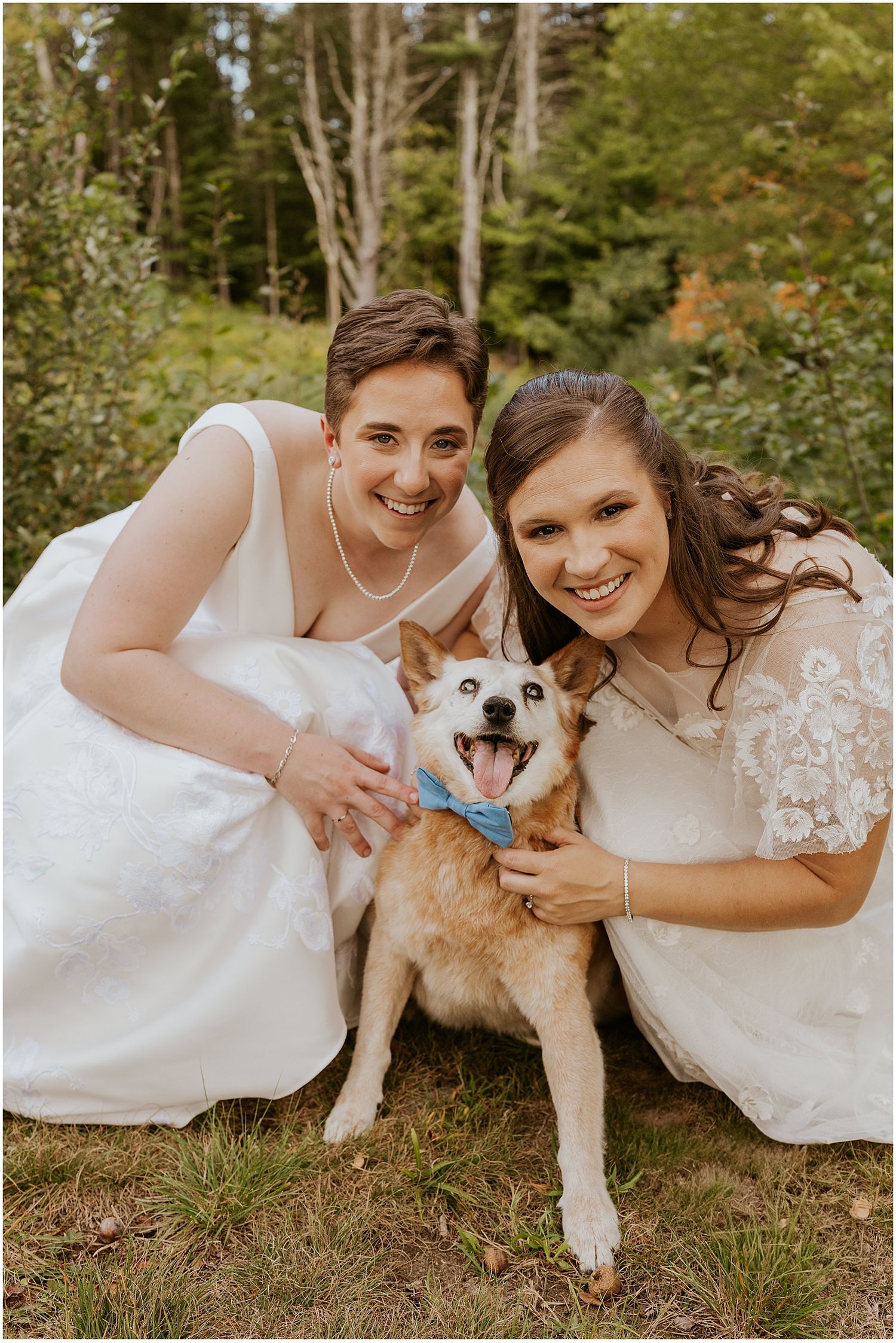 Two people in white wedding dresses sit on grass while petting a fluffy white dog during an outdoor photoshoot.