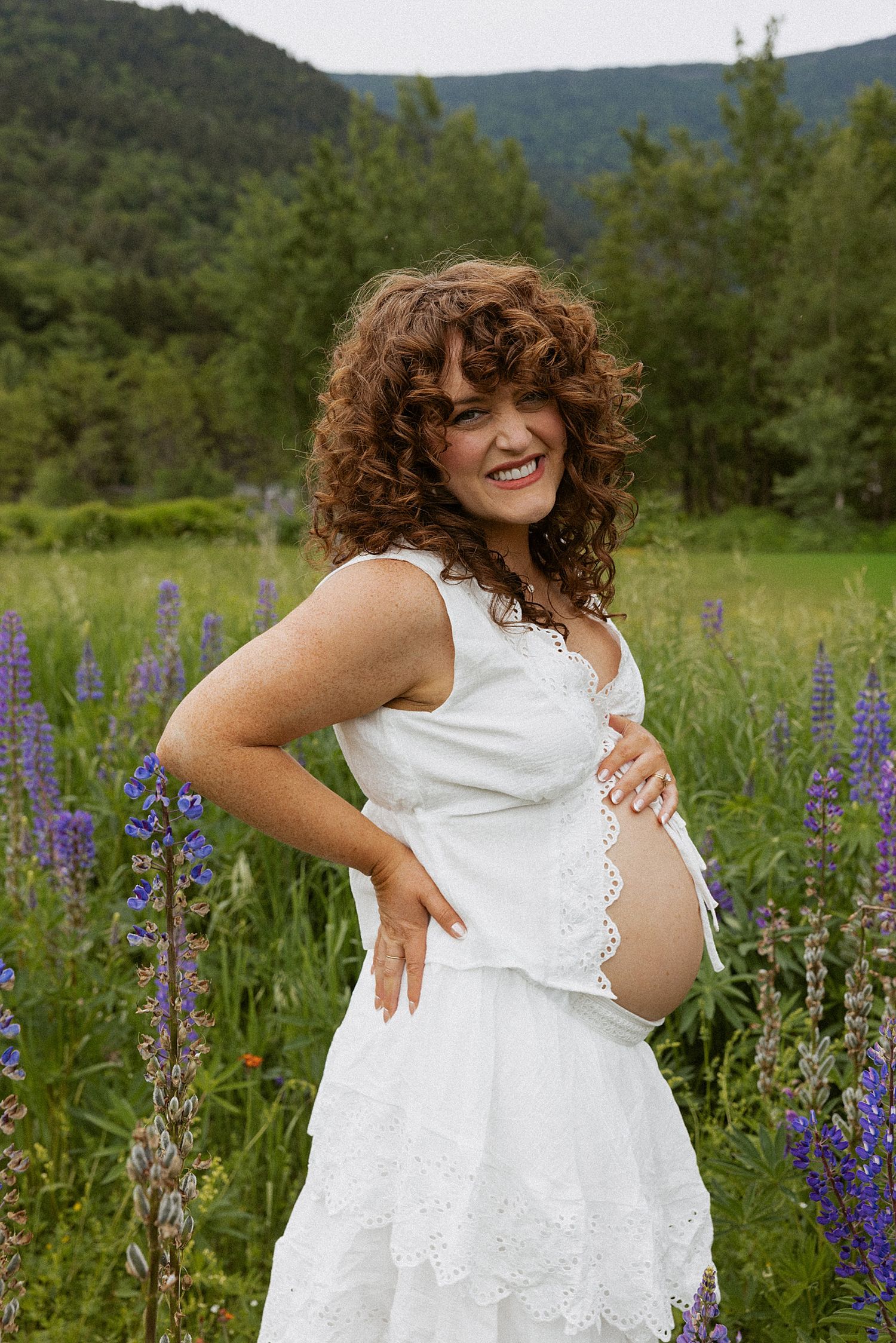 Pregnant woman in white dress holding baby bump in purple lupine flower field with mountains in New Hampshire, taking motherhood photos