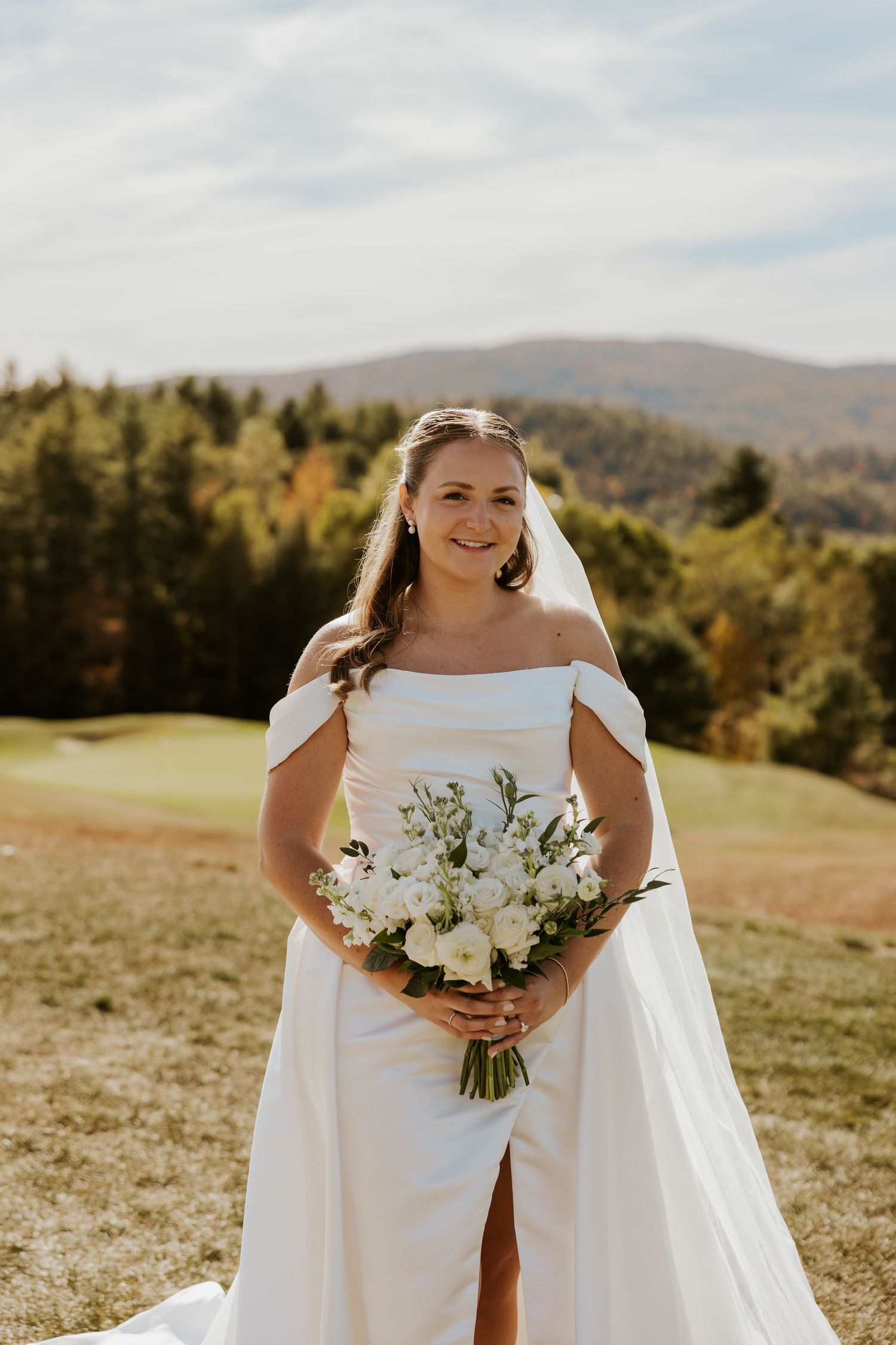Bride in off-shoulder white gown holding white floral bouquet with mountain landscape background.