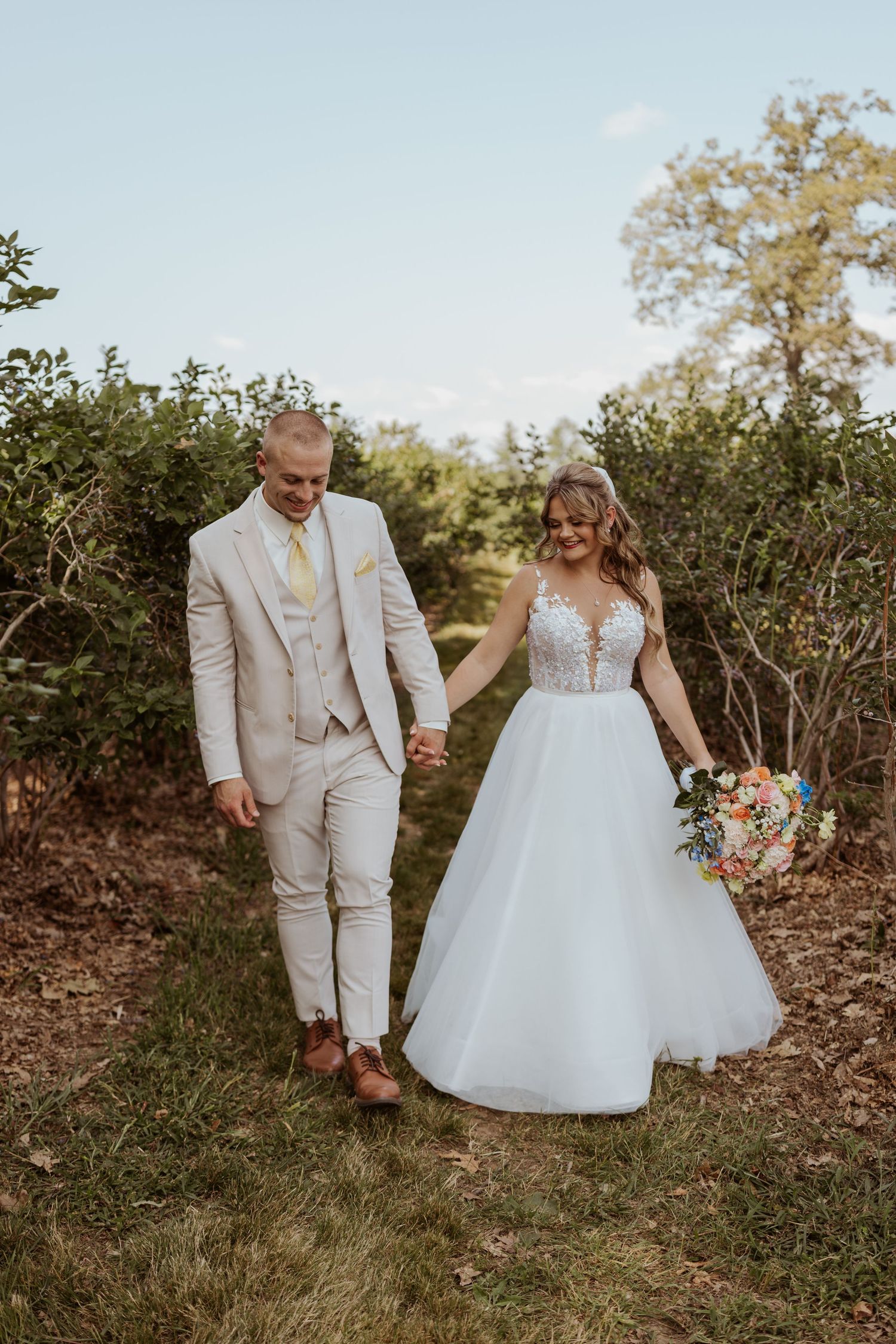 Bride and groom walk hand in hand through an orchard, bride carrying a vibrant floral bouquet.
