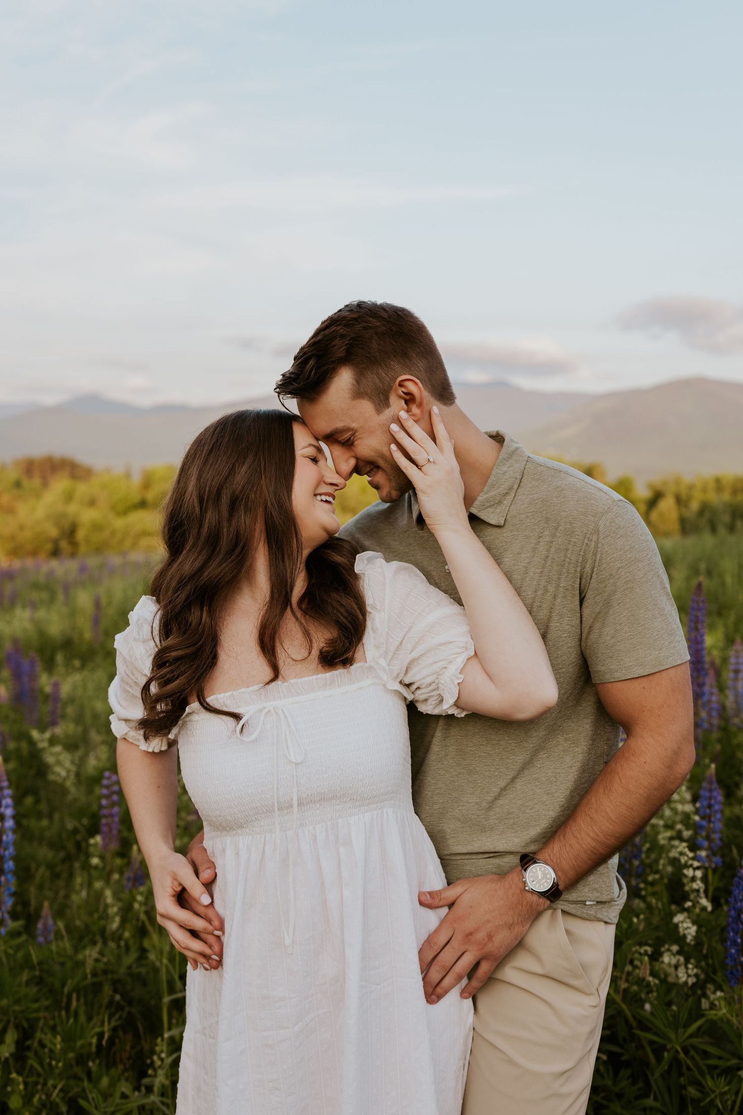 Couple sharing a tender forehead kiss surrounded by blooming purple lupines and mountains.