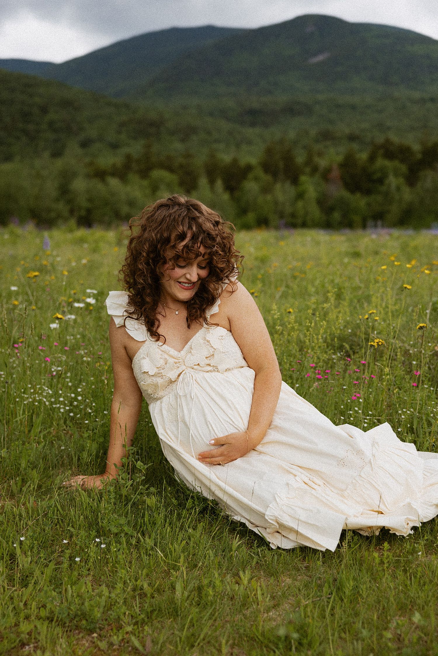 Pregnant woman in white dress sitting in wildflower meadow in New Hampshire's White Mountains taking motherhood photos