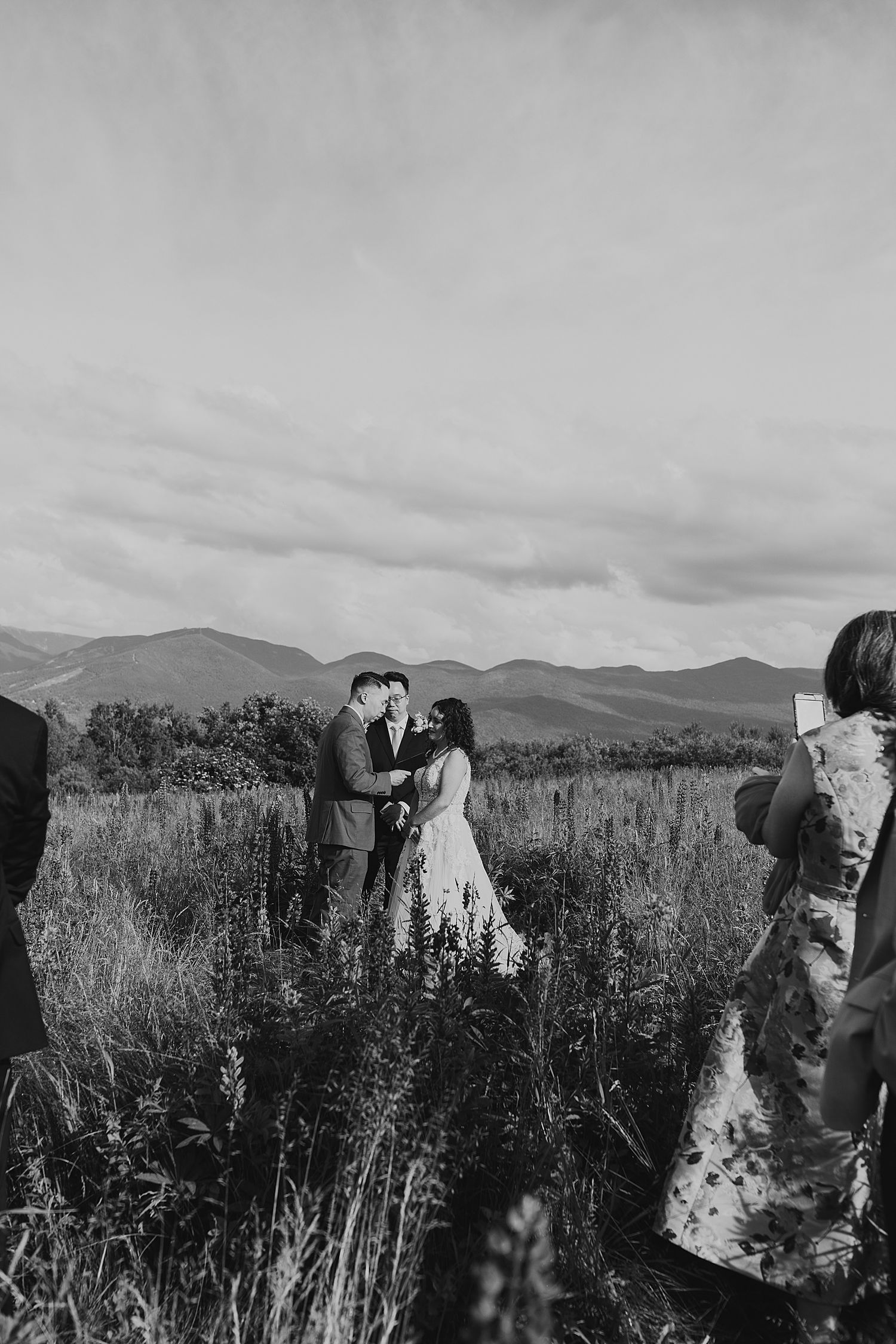 Black and white photo of a couple exchanging vows in a mountain meadow with guests watching.