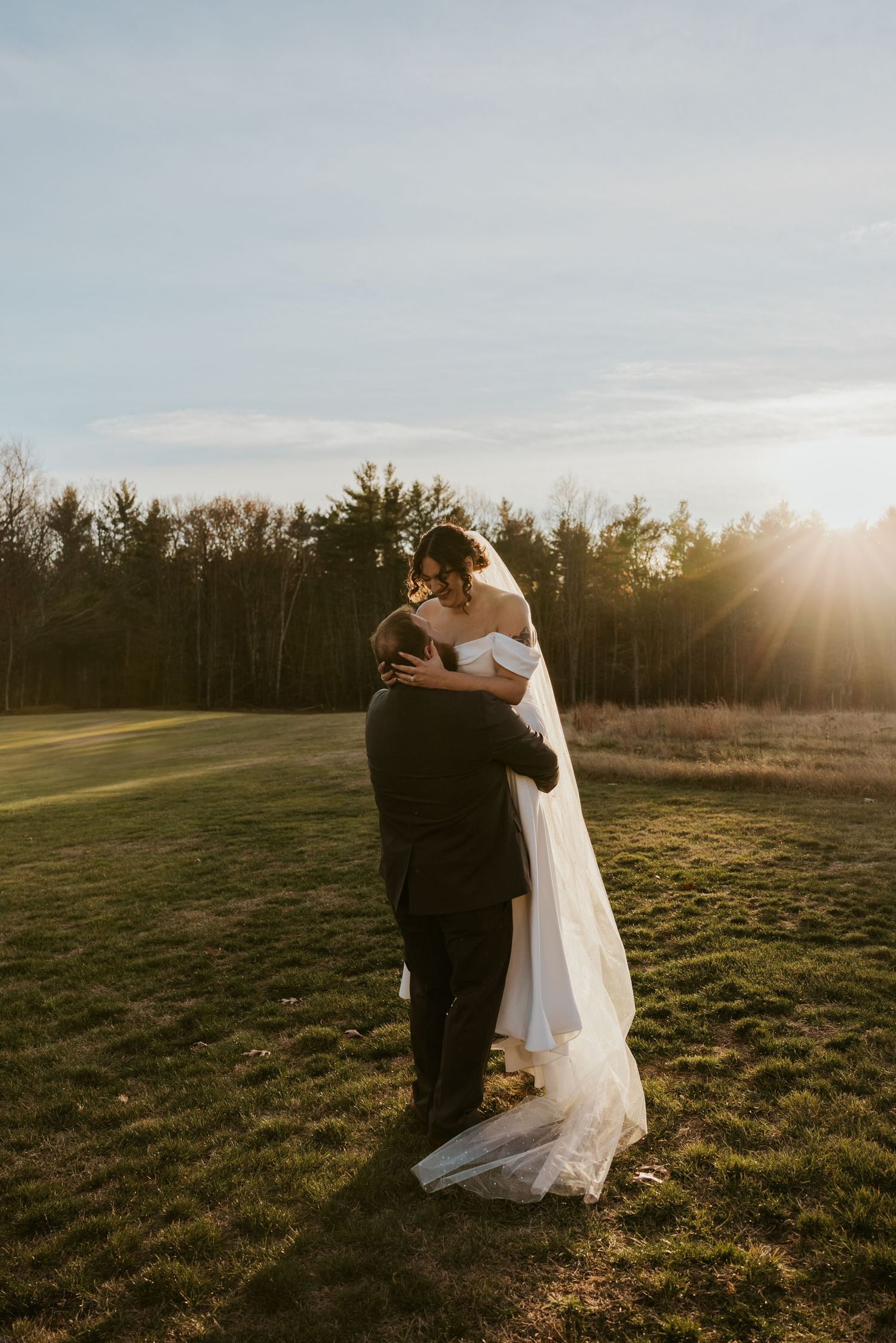 Bride in off-shoulder gown and groom in black suit embrace at golden hour on an open grassy field.