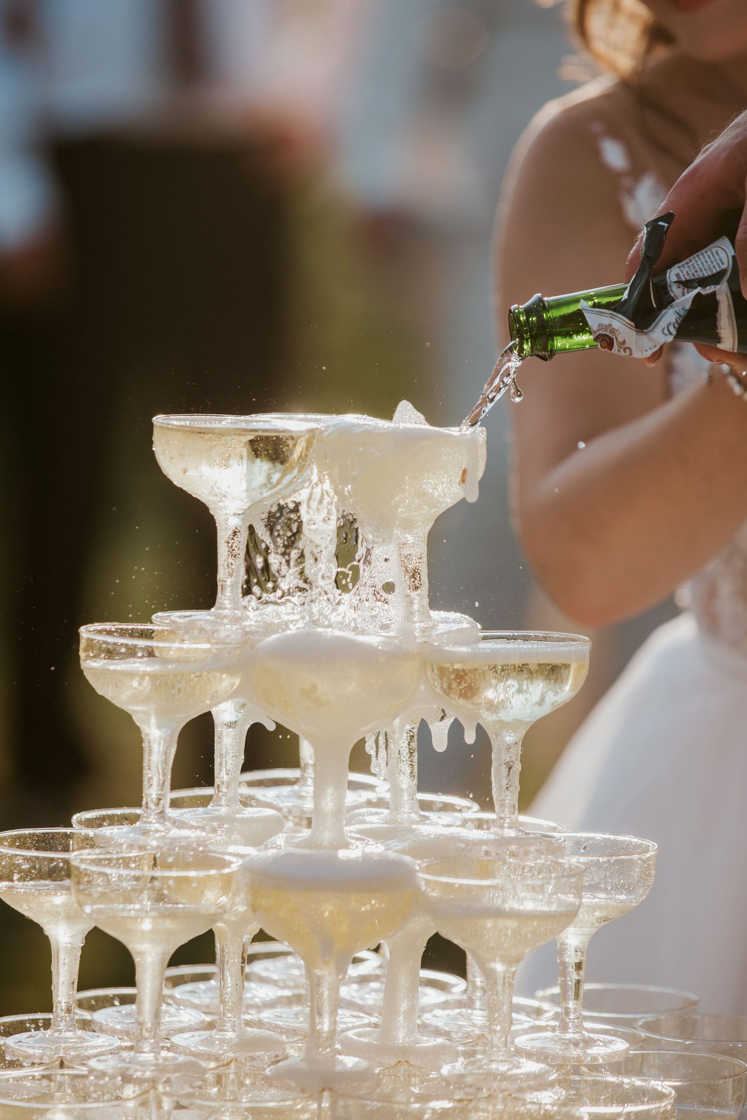 Champagne overflowing from a pyramid tower of coupe glasses at an outdoor wedding.