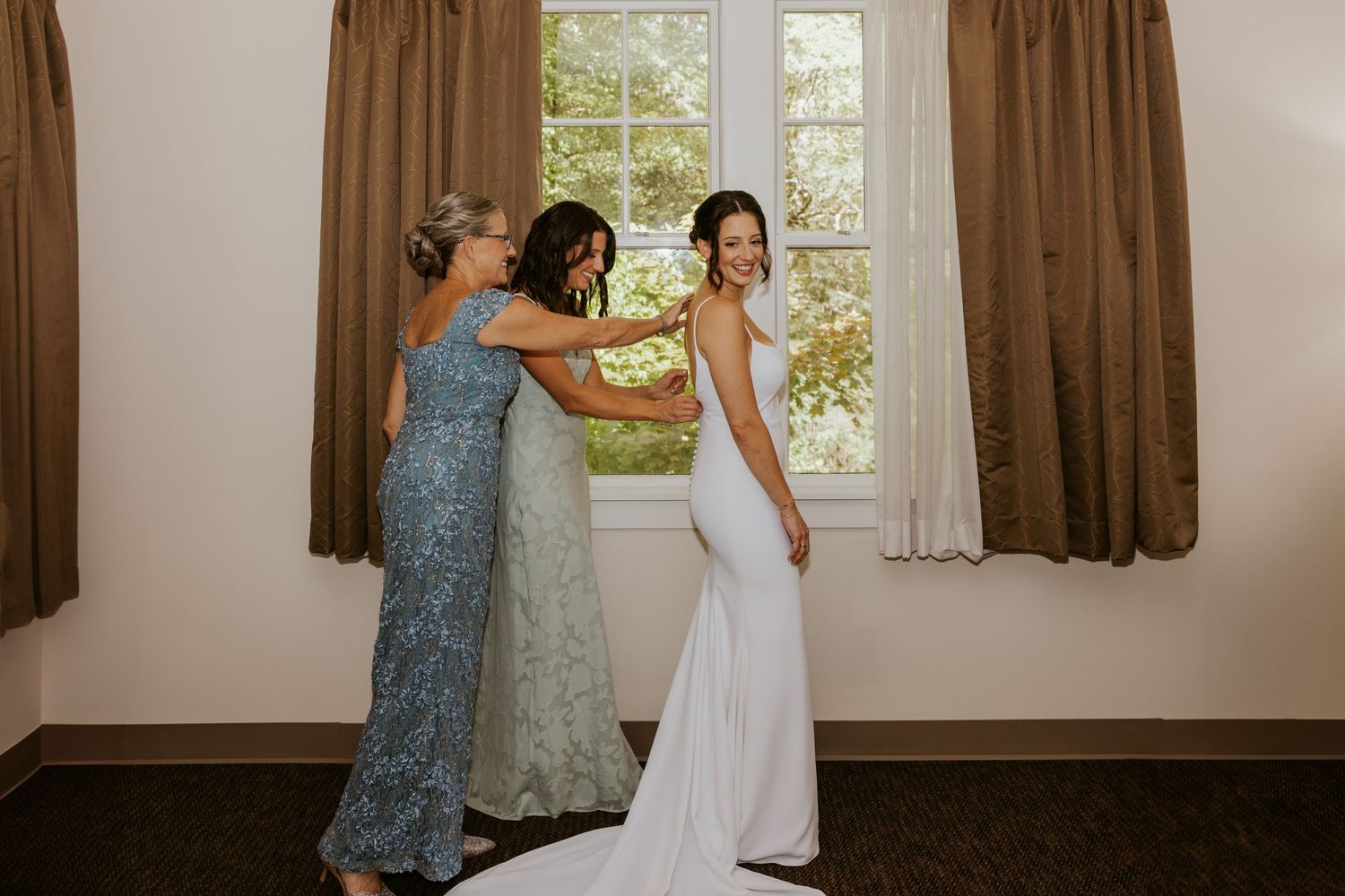 Two women helping bride into her white wedding gown in a sunlit hotel room before the ceremony.