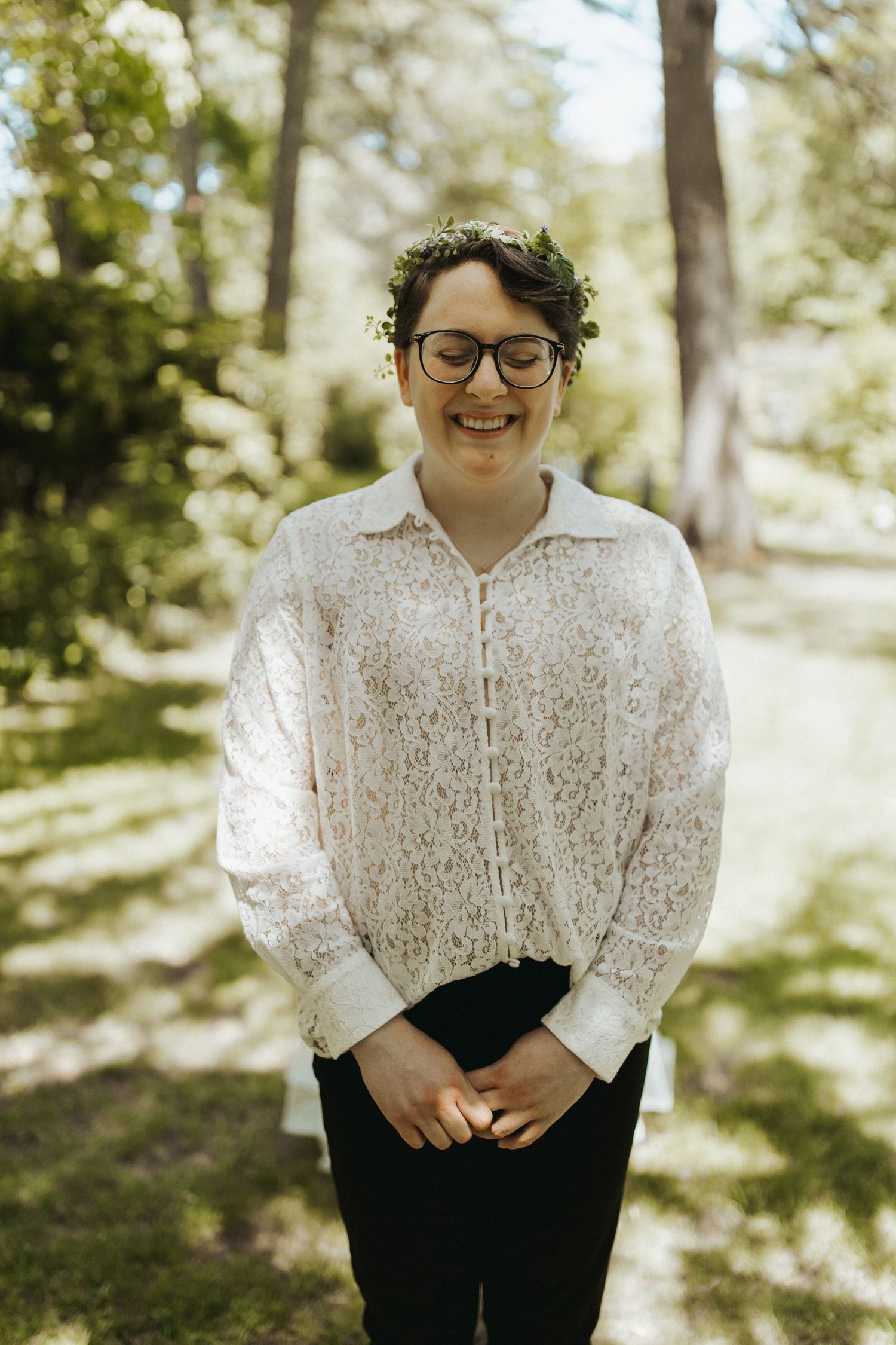 Portrait in white lace top and black pants against a sunlit natural backdrop.