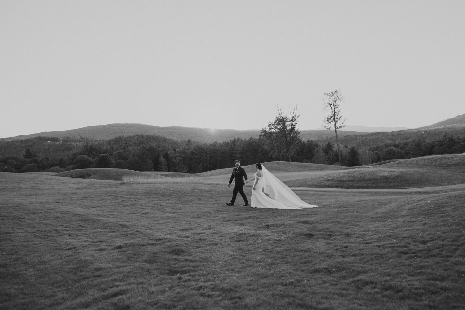 Black and white photo of bride and groom walking hand in hand across open field with rolling hills backdrop.