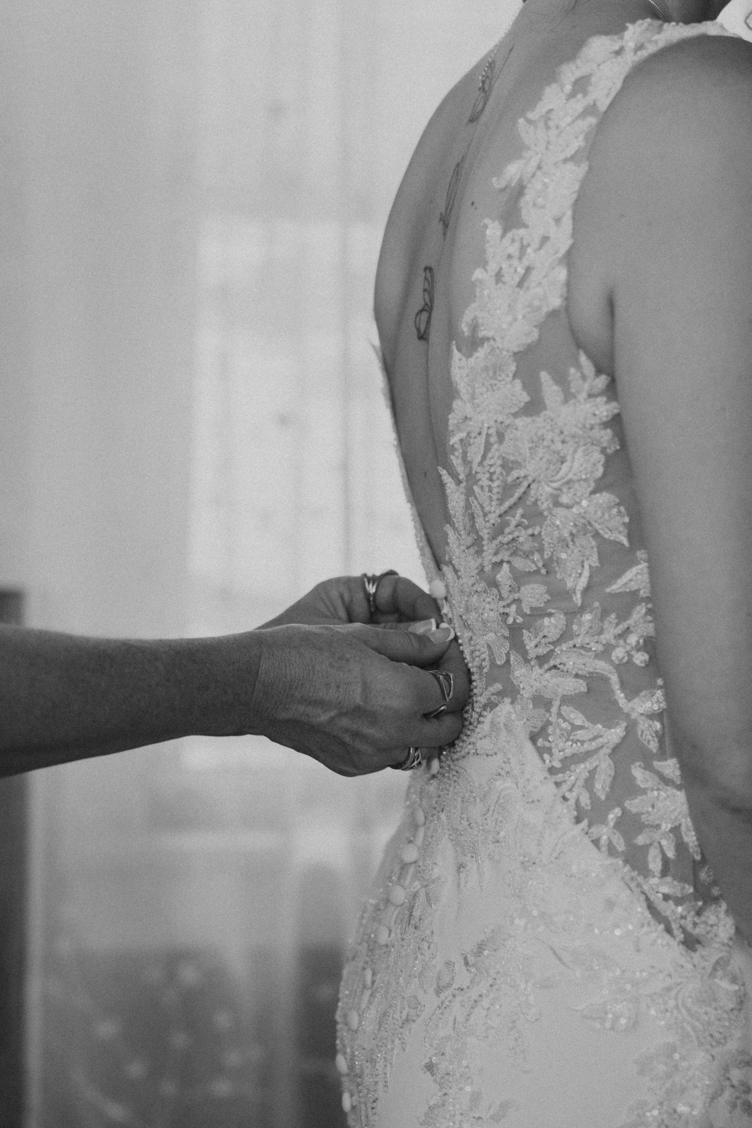 Black and white photo of hands fastening buttons on a lace wedding dress back.