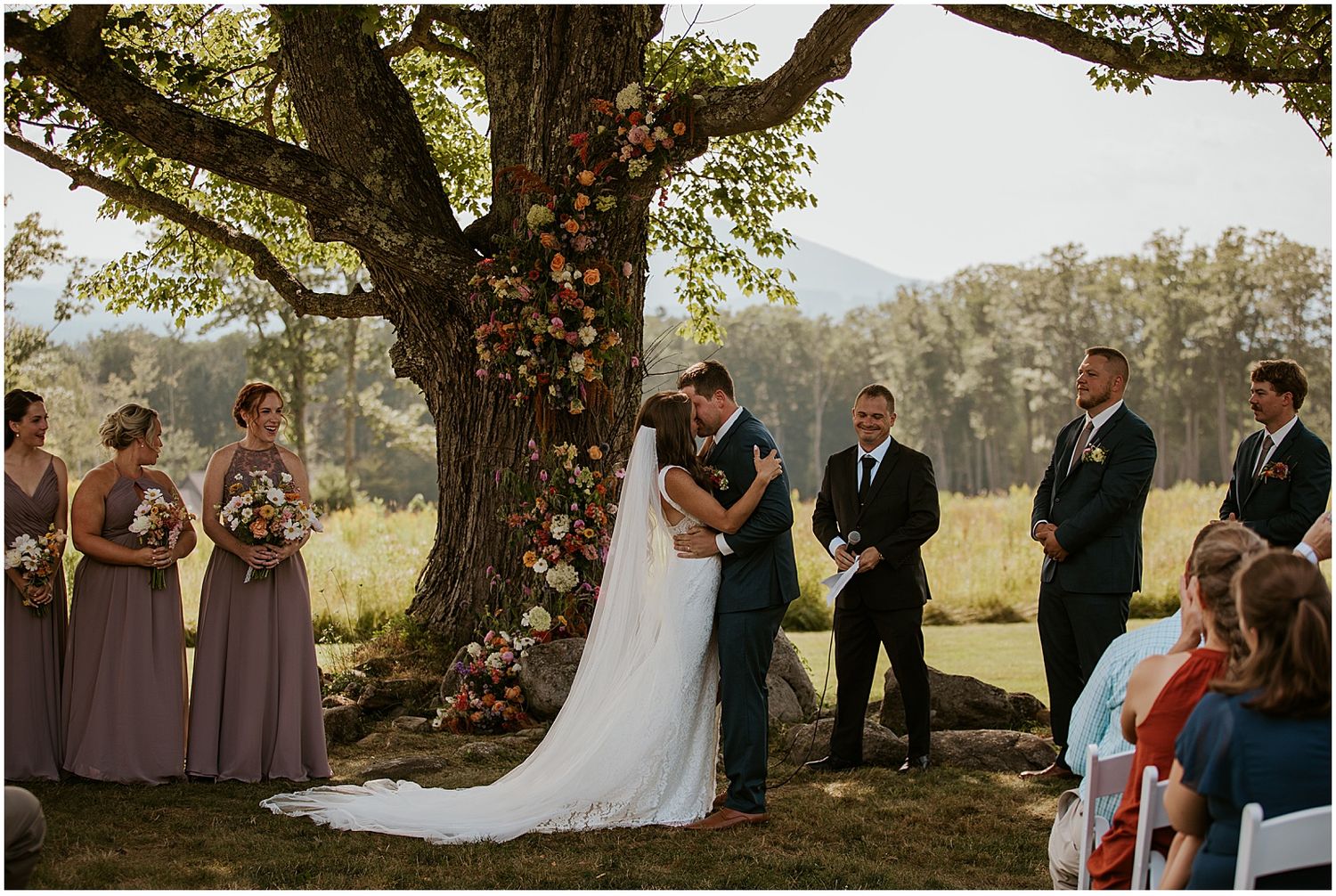 Outdoor wedding ceremony taking place under large tree with guests seated on white chairs.