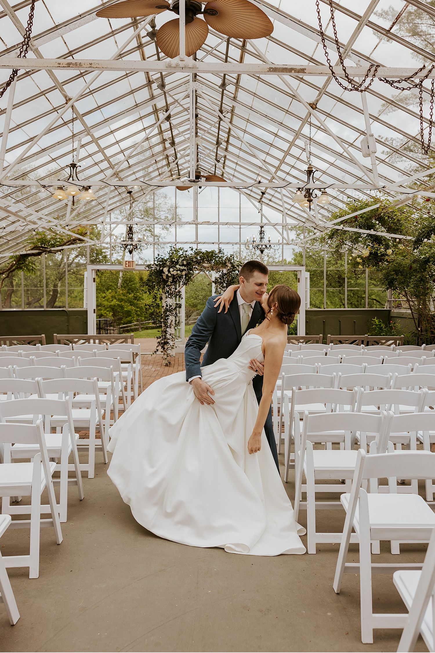 A wedding ceremony setup in a bright greenhouse conservatory with rows of white chairs and natural light streaming in.