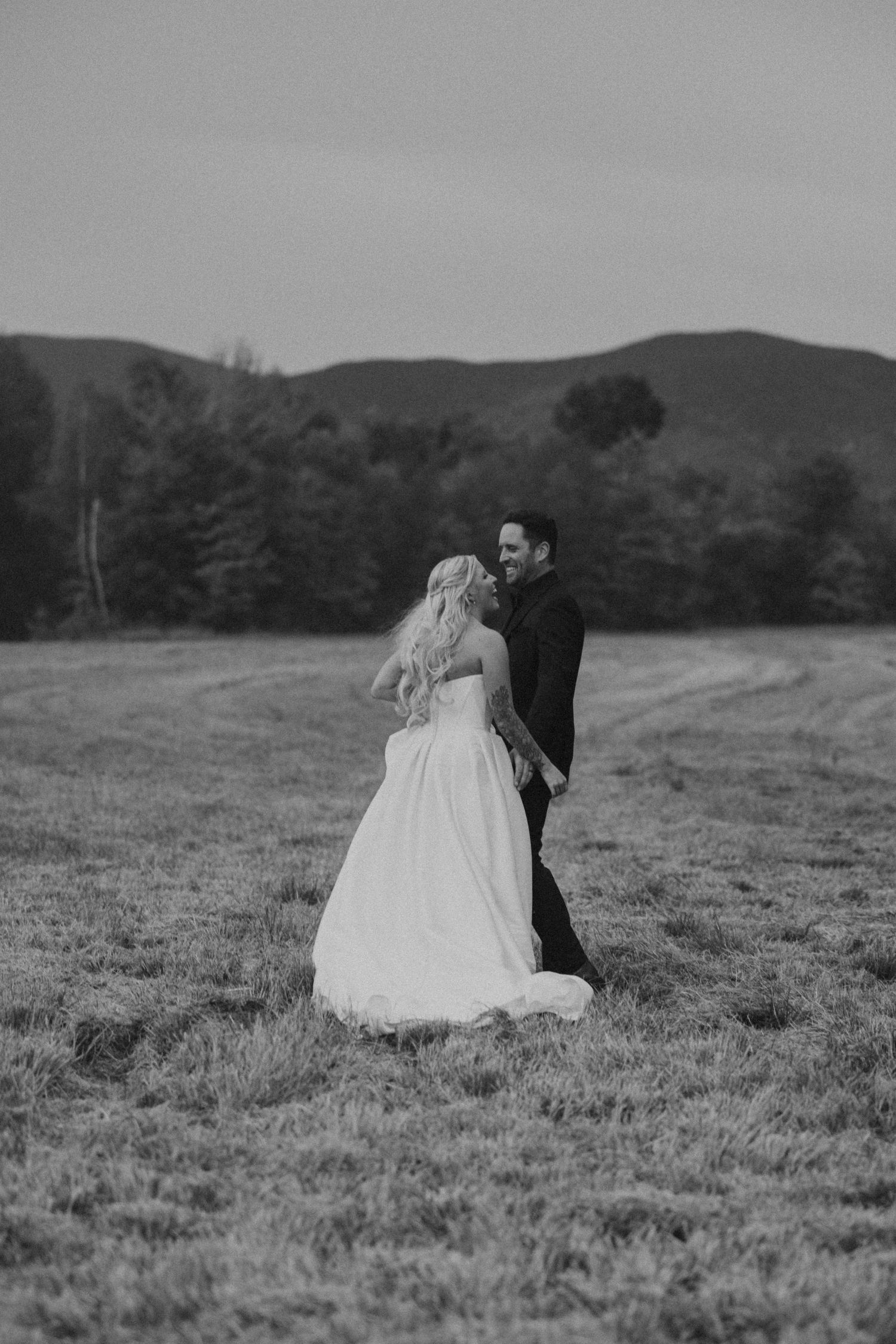 Black and white photo of a bride and groom walking in a field with mountains and trees in the background.