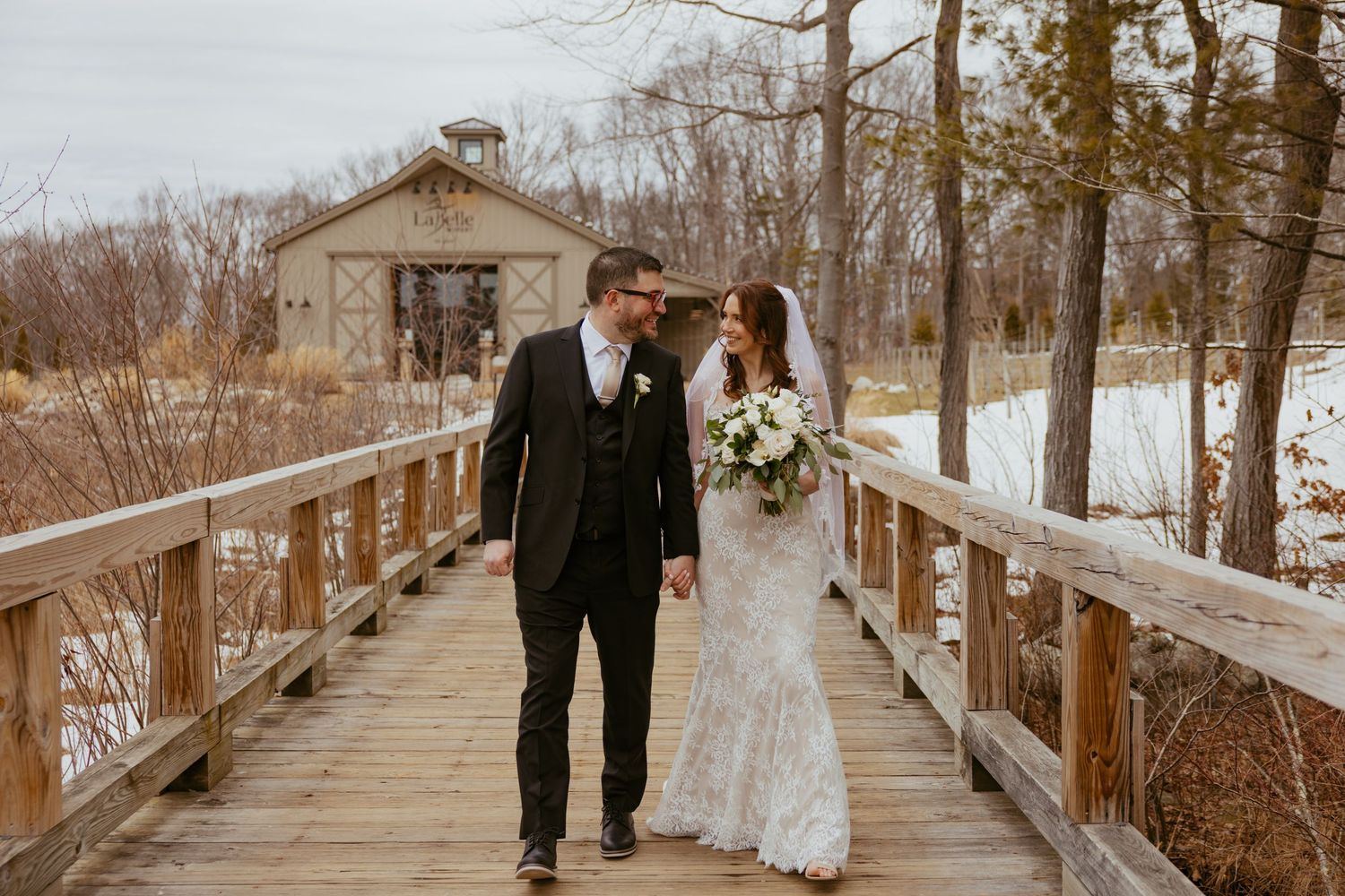 A couple walks along a rustic wooden boardwalk surrounded by bare winter trees in a romantic outdoor setting.