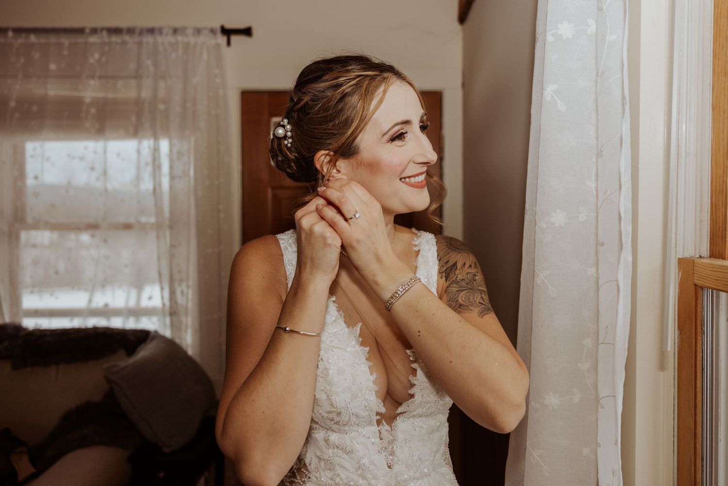Bride smiling while putting on earrings in a softly lit room on her wedding day.