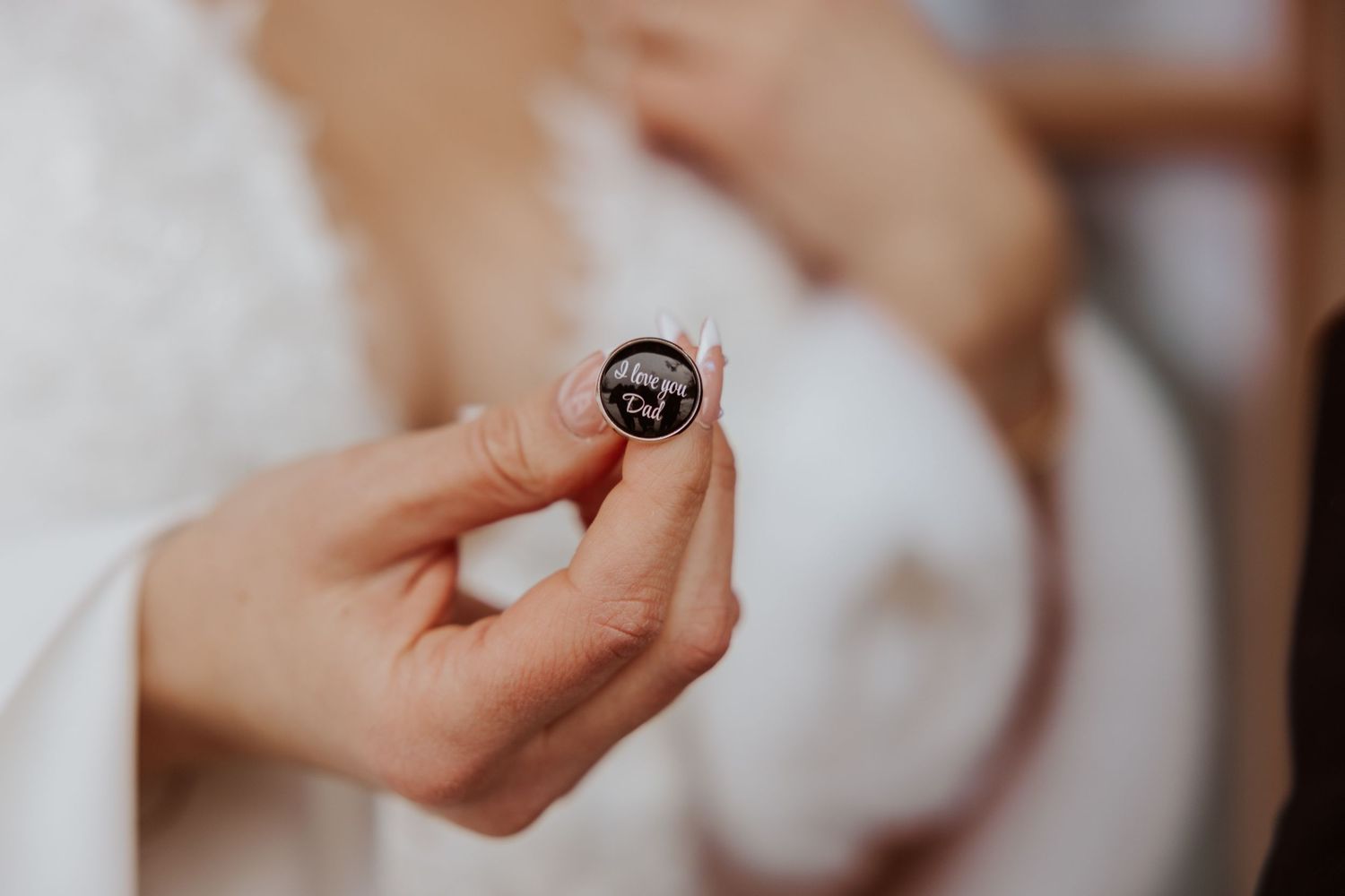 Close-up of a bride holding a small round wedding cufflink with an engraved design on her wedding day.