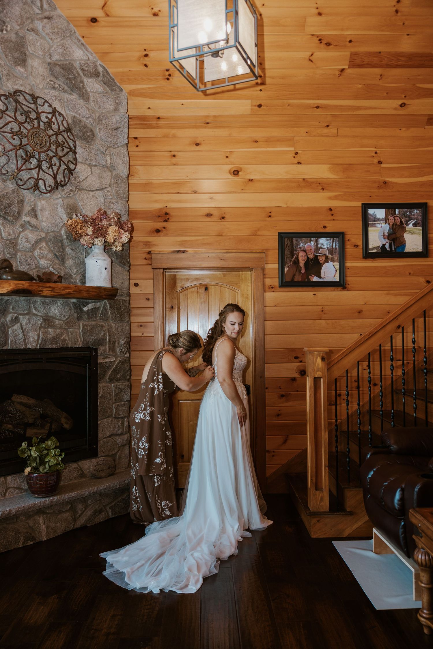 A wedding dress fitting in a rustic cabin interior with wooden walls and stone fireplace.