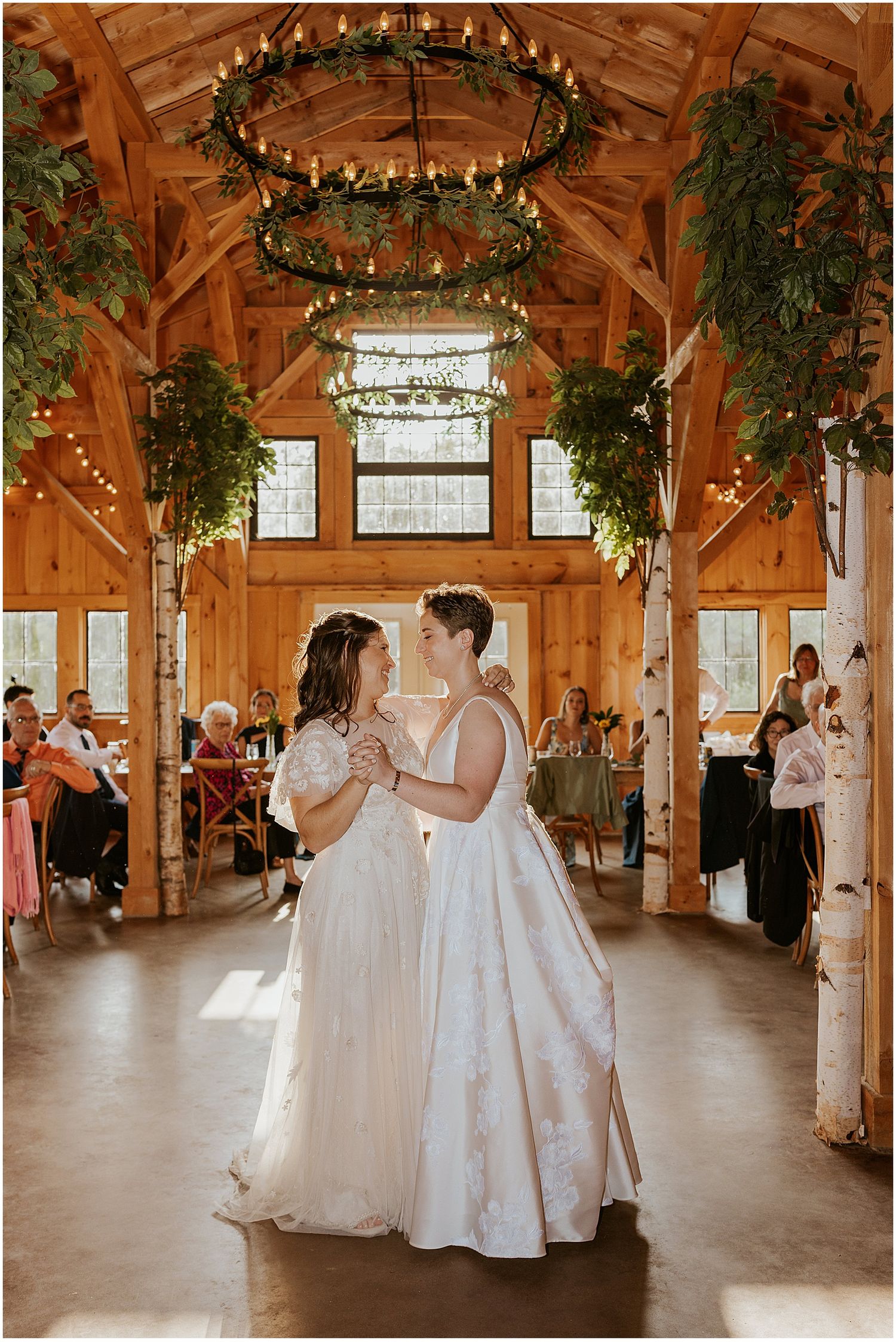 A rustic barn wedding venue with wooden beams, twinkle lights, and birch trees, while brides share their first dance