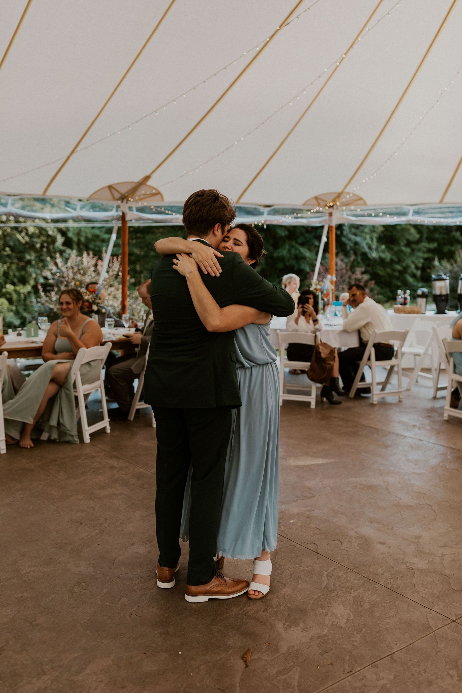Wedding party members dancing together under a white tent during reception celebrations.