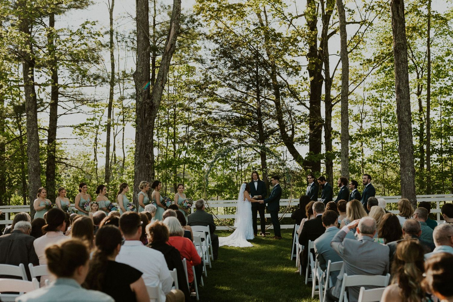Chrissy and Mike holding hands as their wedding ceremony begins. It is a lovely spring day with the sun shining.
