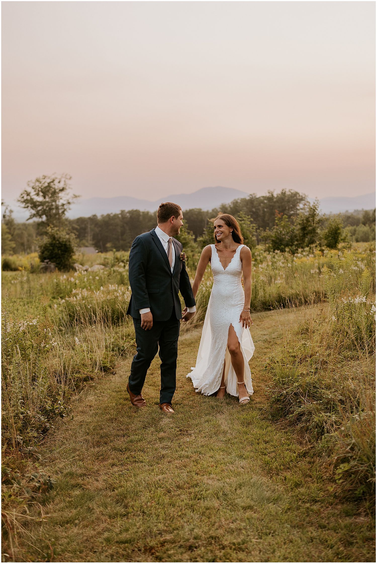 Couple walking through a meadow path during sunset with mountains in the background.