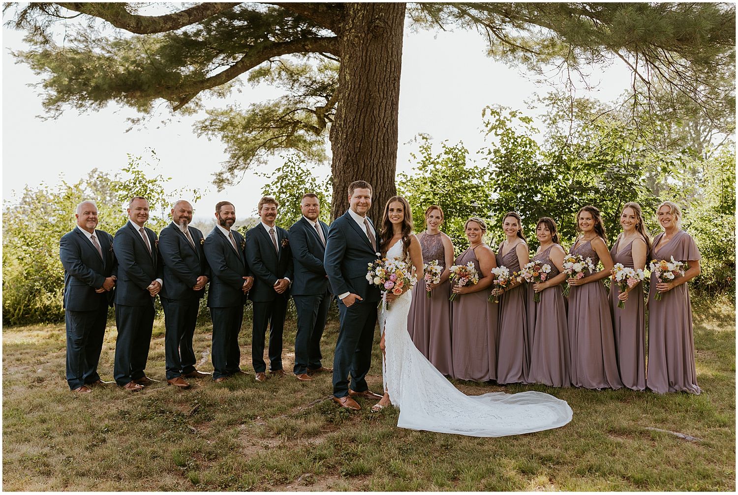 Wedding party in purple dresses and suits lined up outdoors under large tree.