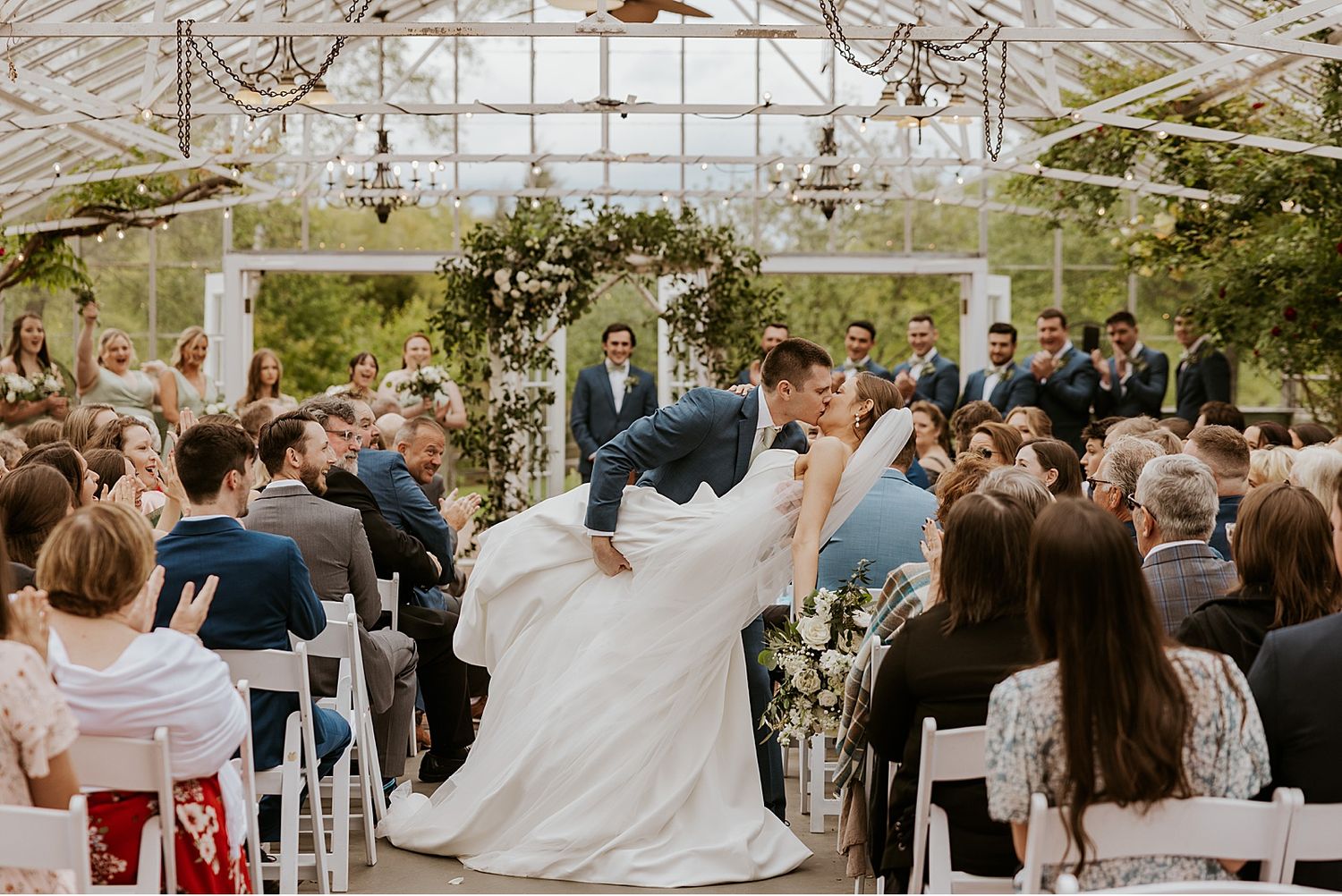 Wedding celebration inside a sunlit greenhouse with guests seated in rows.