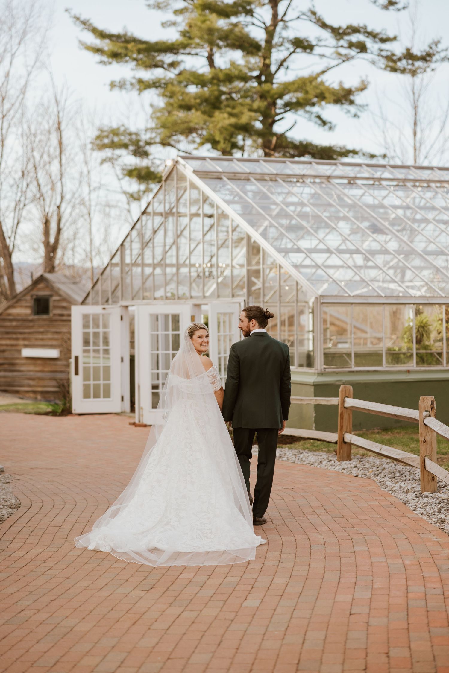 Bride and groom walk hand in hand on brick path before glass greenhouse wedding venue.