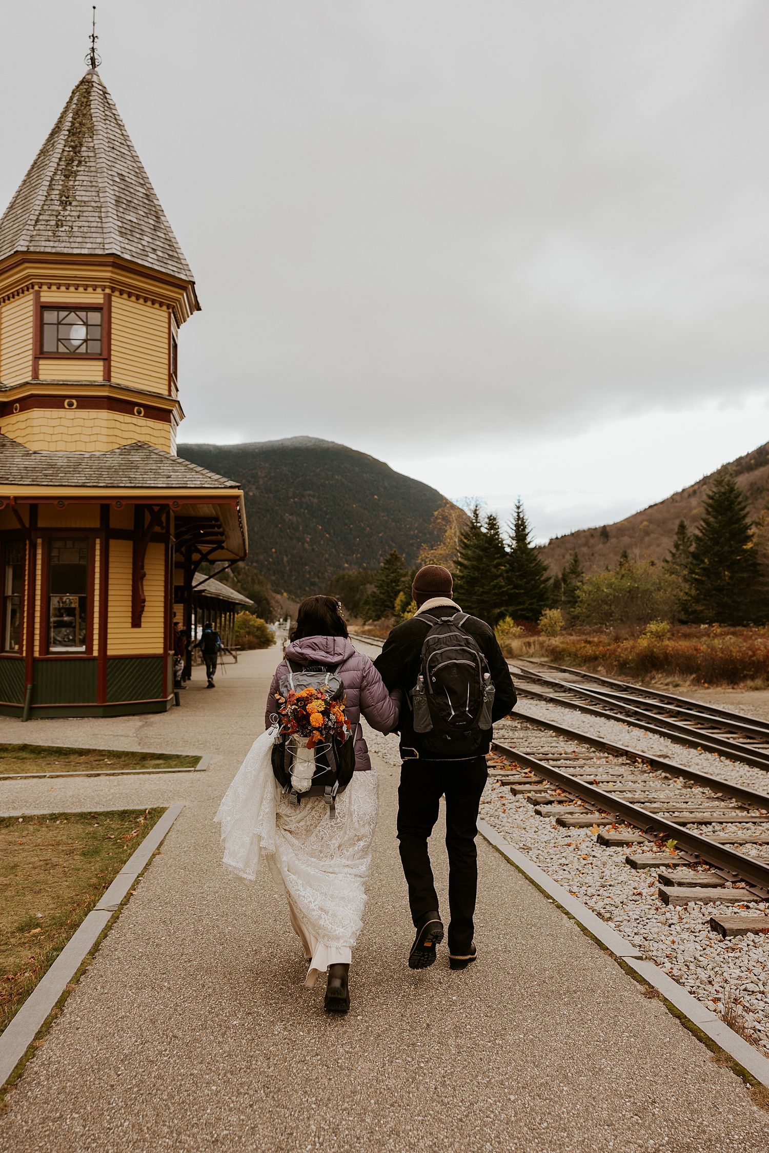 Bride and groom with backpacks walk along a mountain train station platform after their wedding.