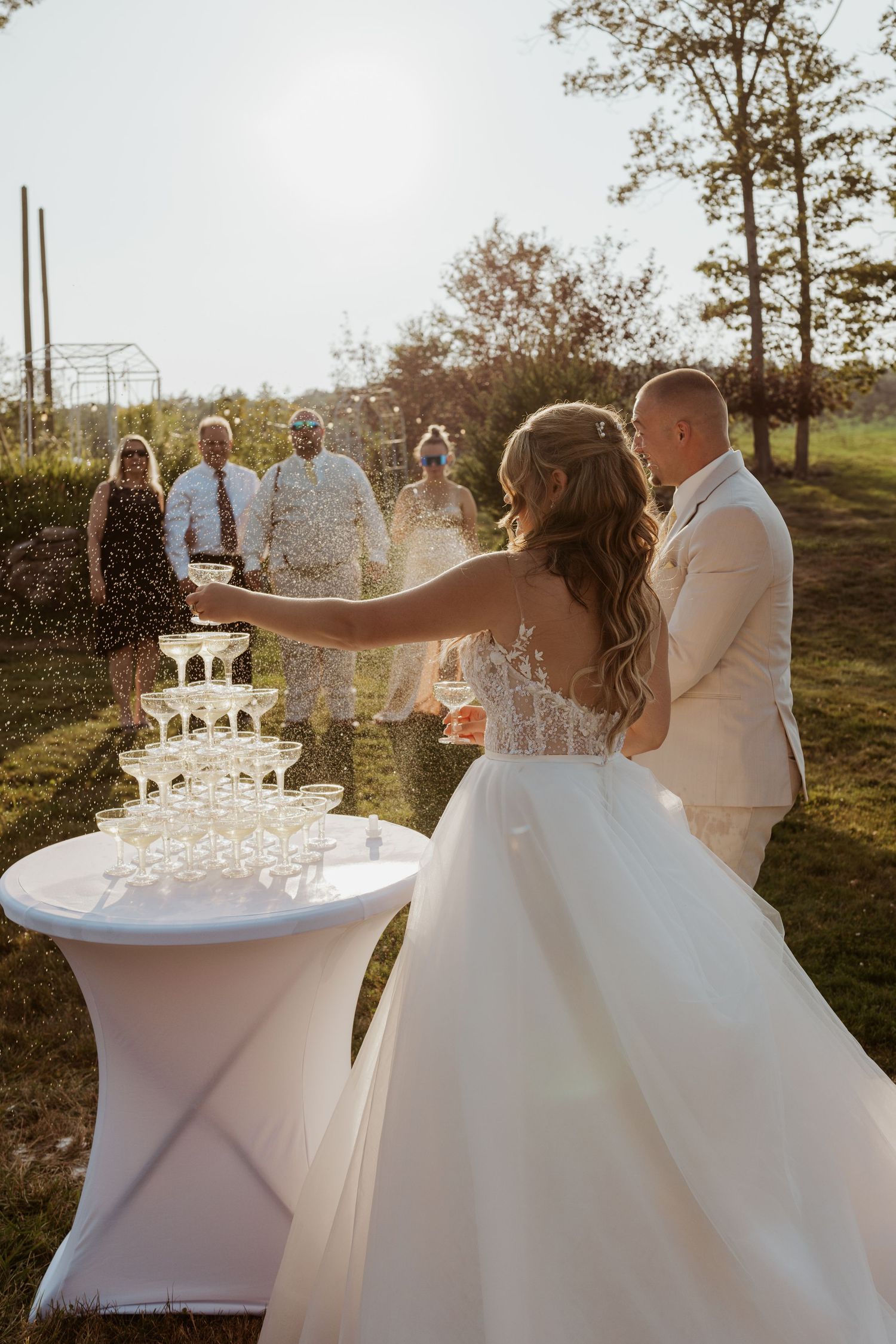 Bride and groom spraying champagne tower with wedding guests watching in sunny outdoor setting.