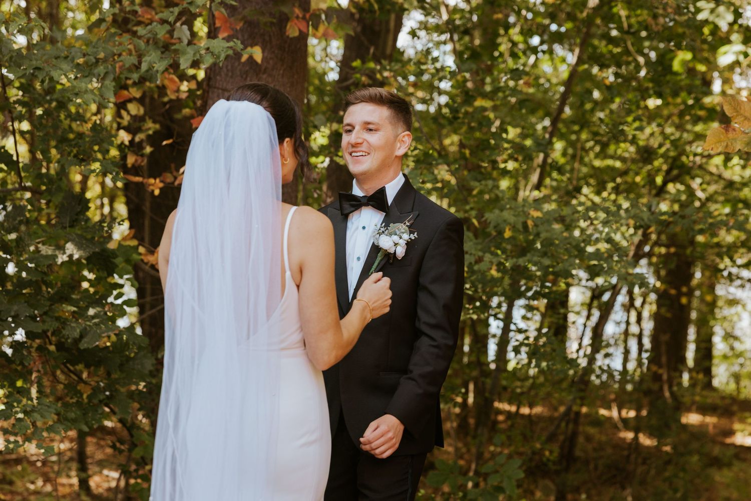 Groom in black tuxedo smiling at bride during outdoor first look among autumn forest trees.
