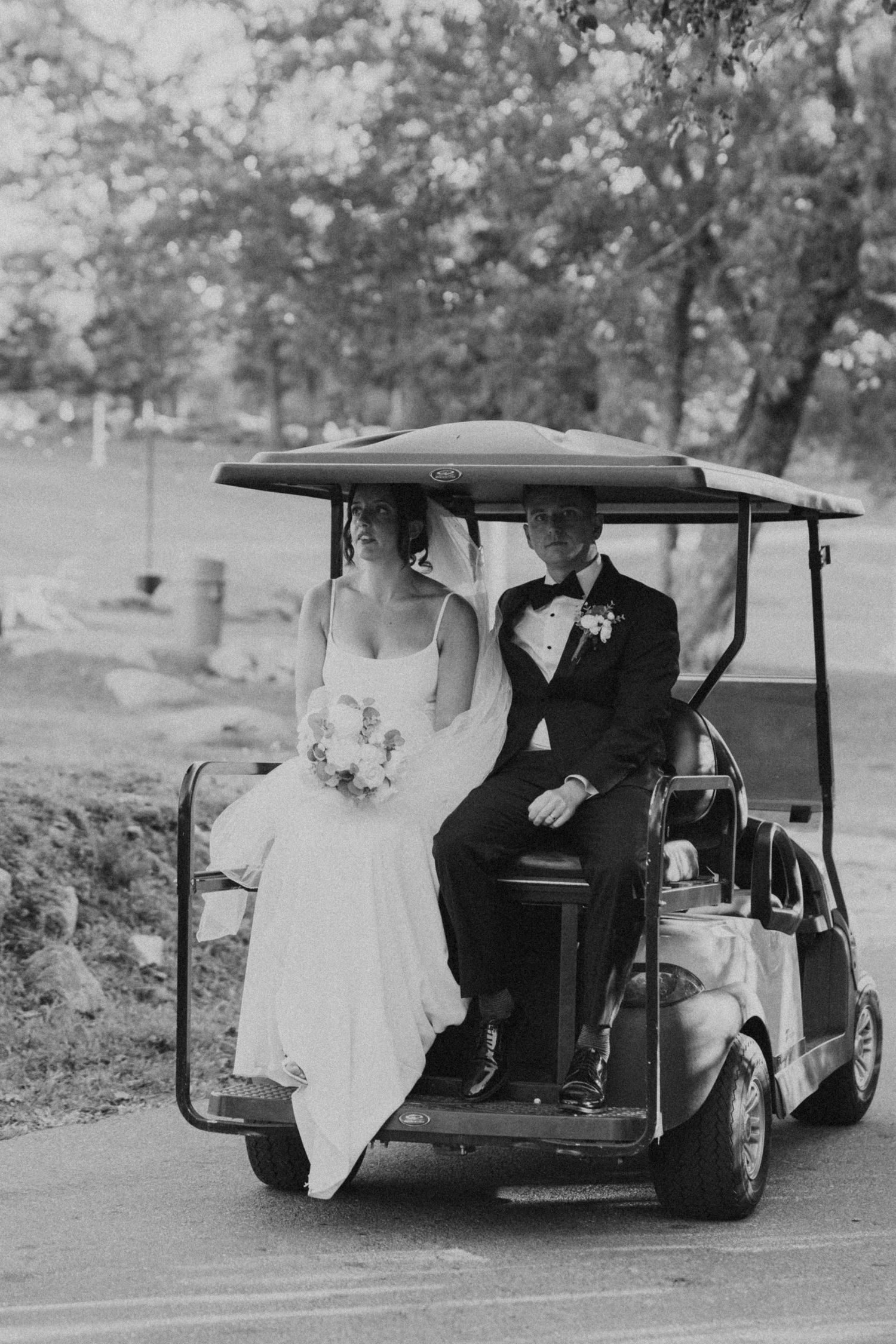 Bride and groom ride golf cart together in black and white photo at outdoor wedding venue.