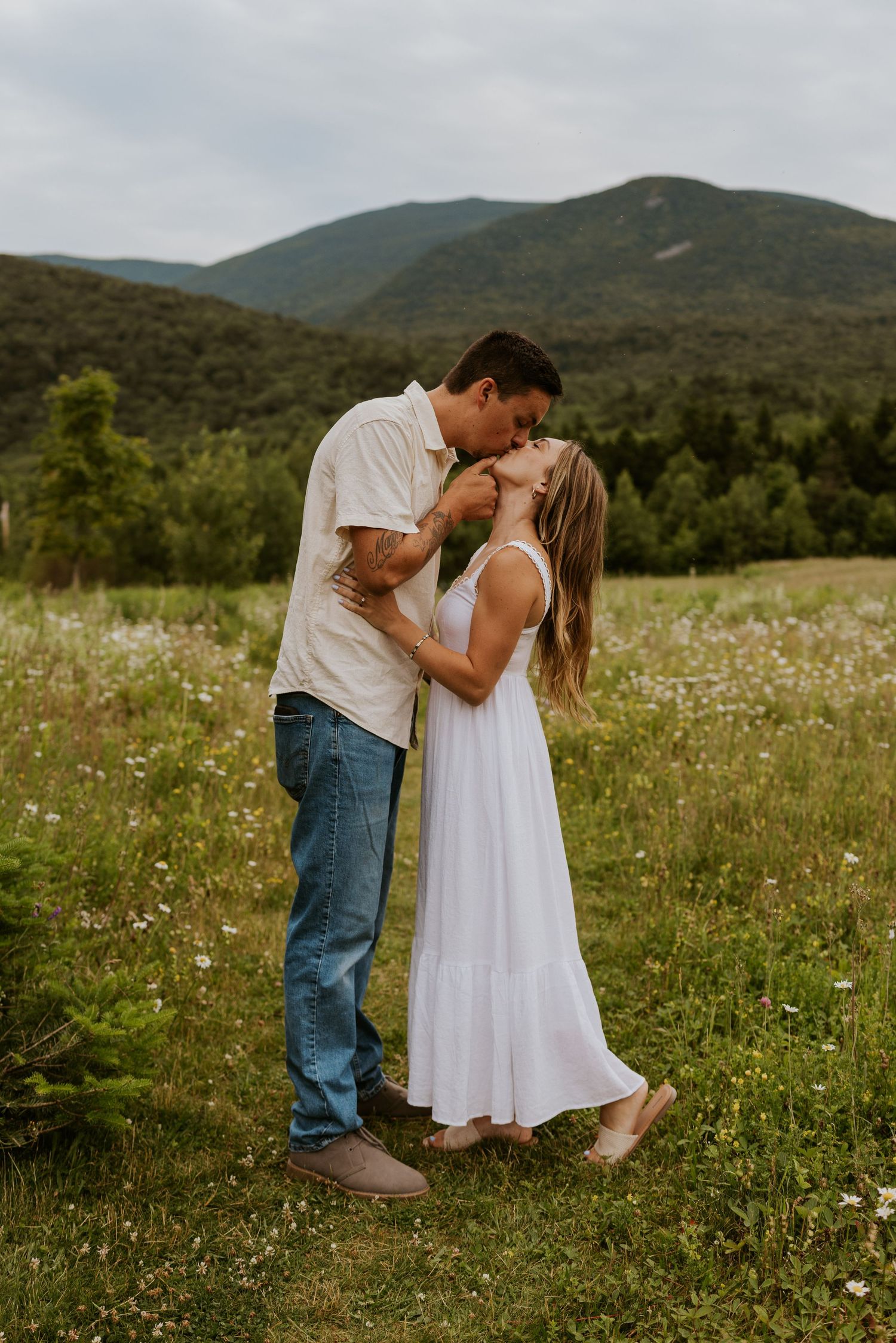 Couple sharing a tender kiss in lush green field with rolling mountains in the background.