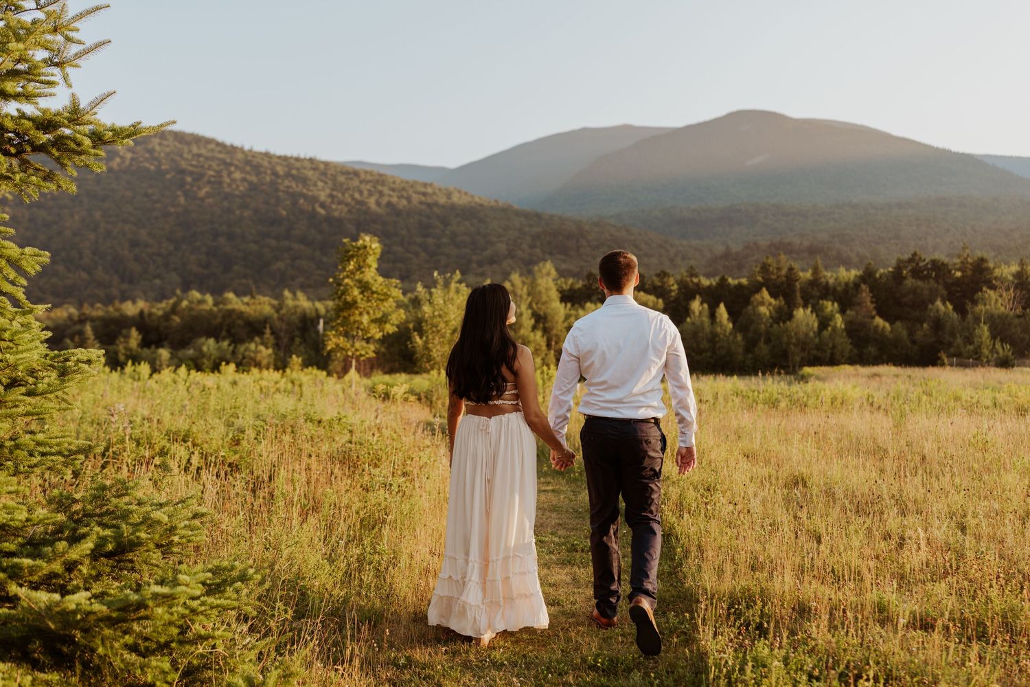 Couple walking hand in hand through a sunlit meadow with stunning mountain views ahead.