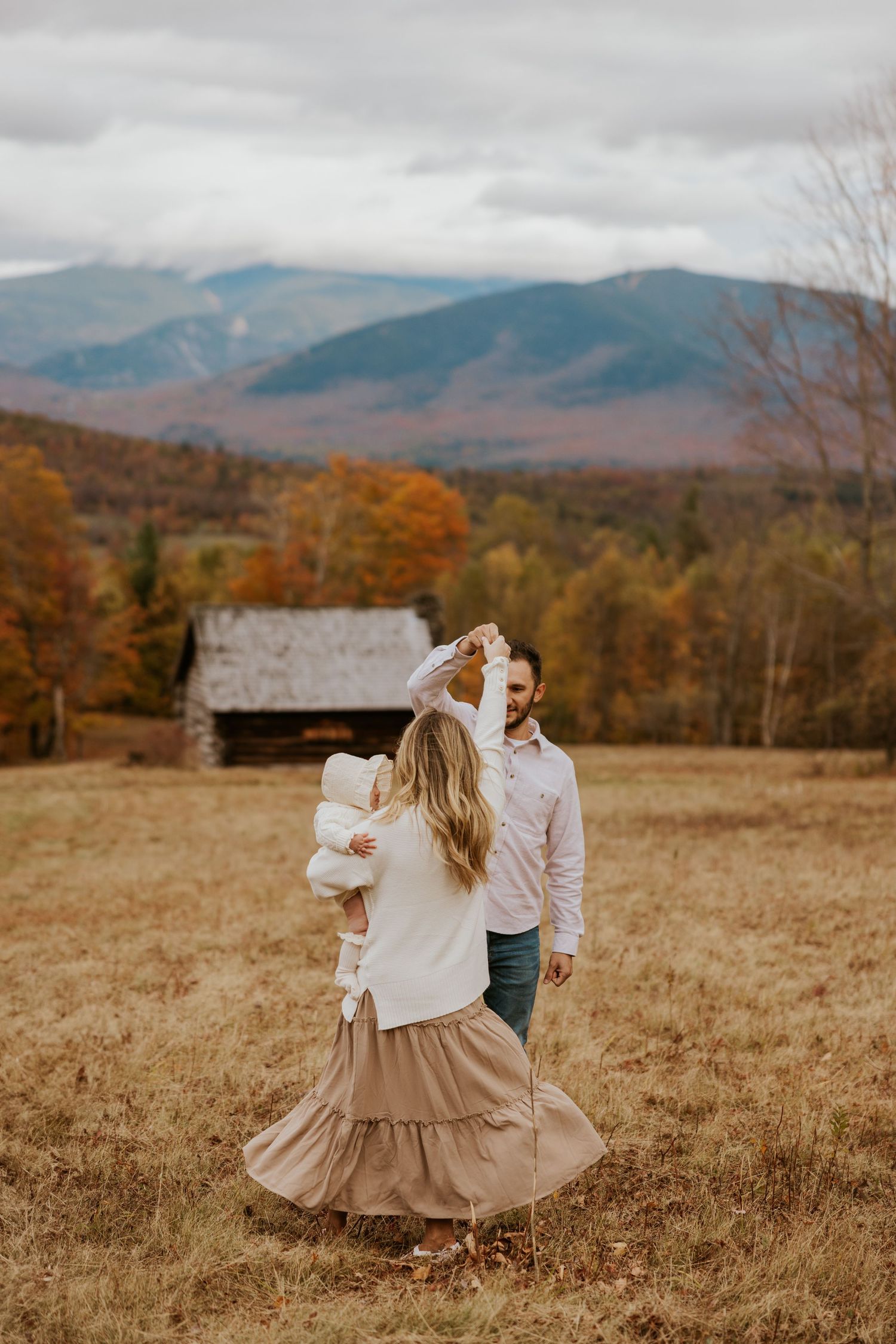 Couple dancing in golden autumn field with mountains and barn backdrop, mother holding newborn baby.