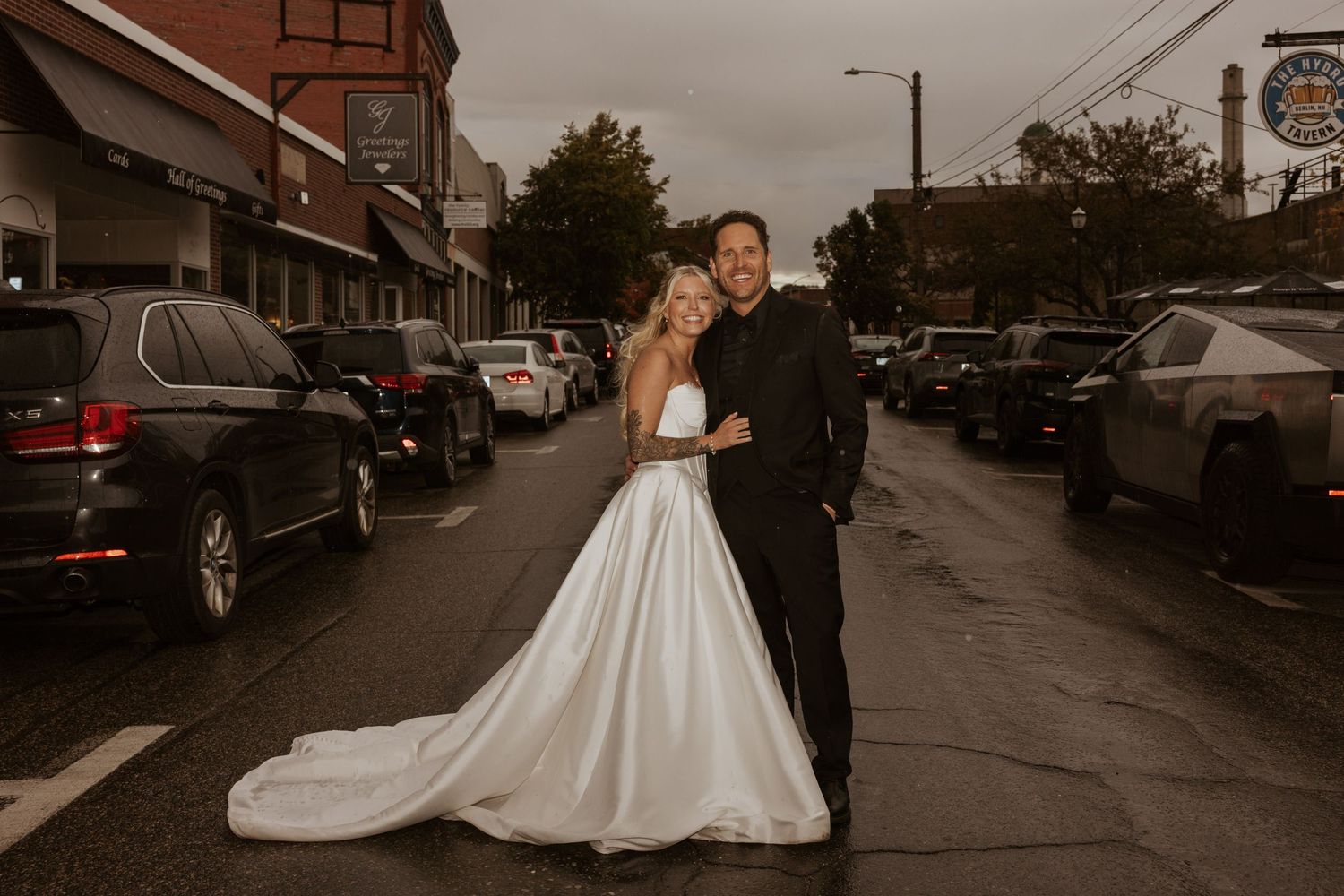Bride and groom posing together on a city street in wedding attire with cars and storefronts behind them.