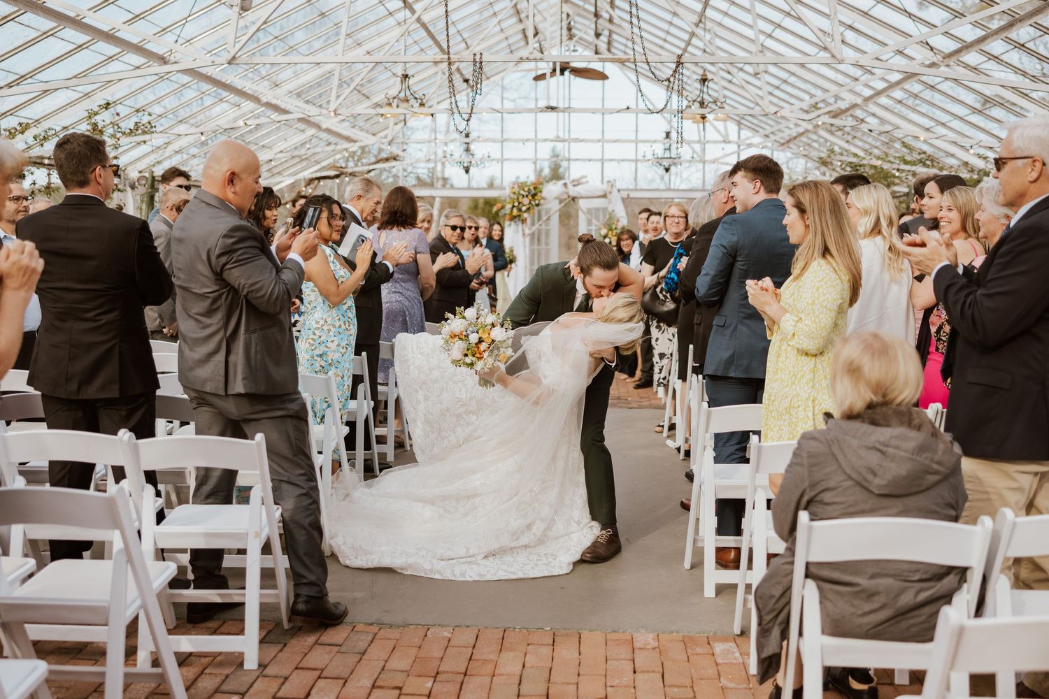 Bride and groom share a romantic dip kiss at their greenhouse wedding ceremony surrounded by applauding guests.