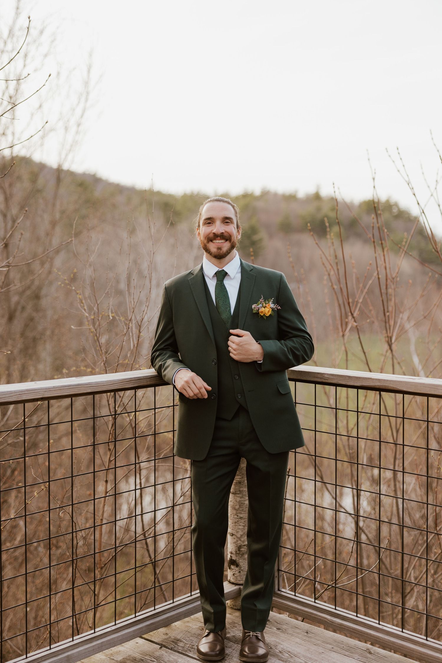 Groom in dark green three-piece suit with floral boutonniere smiles on wooden deck with wooded hillside backdrop.
